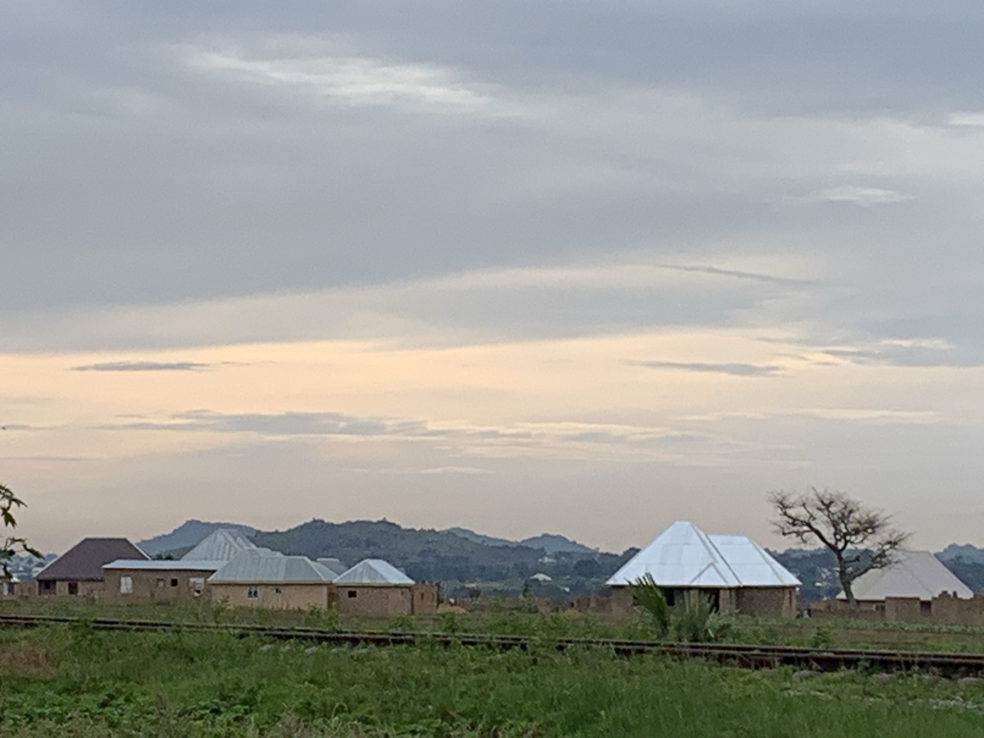 A wide shot captures a pastoral scene at dusk in Kuru Karama, Nigeria. In the foreground, lush green grass and what appear to be railway tracks stretch horizontally across the frame. A line of residential buildings with brick and mud walls and corrugated metal roofs of varying designs forms the middle ground. These homes are varied in size and construction, with some featuring simple rectangular shapes and others with more angular, tent-like roofs. Behind the buildings, rolling hills and mountains form a distant backdrop, their contours softened by the fading light. The sky above is a canvas of soft grays and pale oranges, indicating the transition from day to night, with scattered clouds adding texture. The overall atmosphere is one of quiet rural life at the close of day.