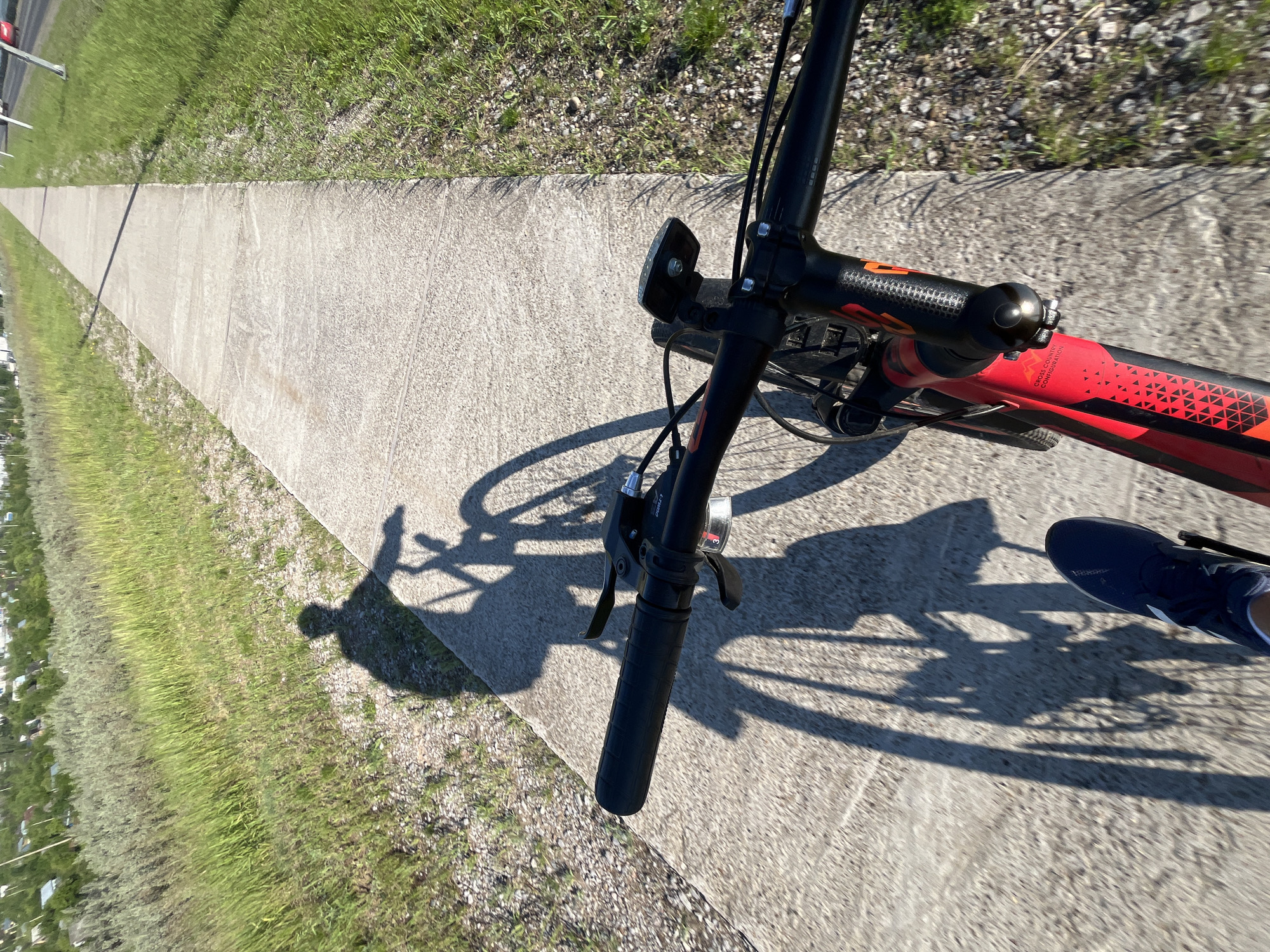 The image shows a first-person perspective of someone riding a red and black mountain bike on a paved multi-use path. The path is bordered by a grassy area with some gravel. In the distance, there are some buildings and trees, suggesting a suburban or semi-rural setting within the city of Chaikovsky, Russia. The sun is casting long shadows from the rider and the bike onto the path, indicating it is either early morning or late afternoon. The rider's left foot, wearing a dark blue athletic shoe, is visible in the lower left corner of the frame. The handlebars of the bike are black with orange accents, and a small black light is attached to the stem. The bike frame has "CROSS COUNTRY CONFIGURATION" written on it in white text.