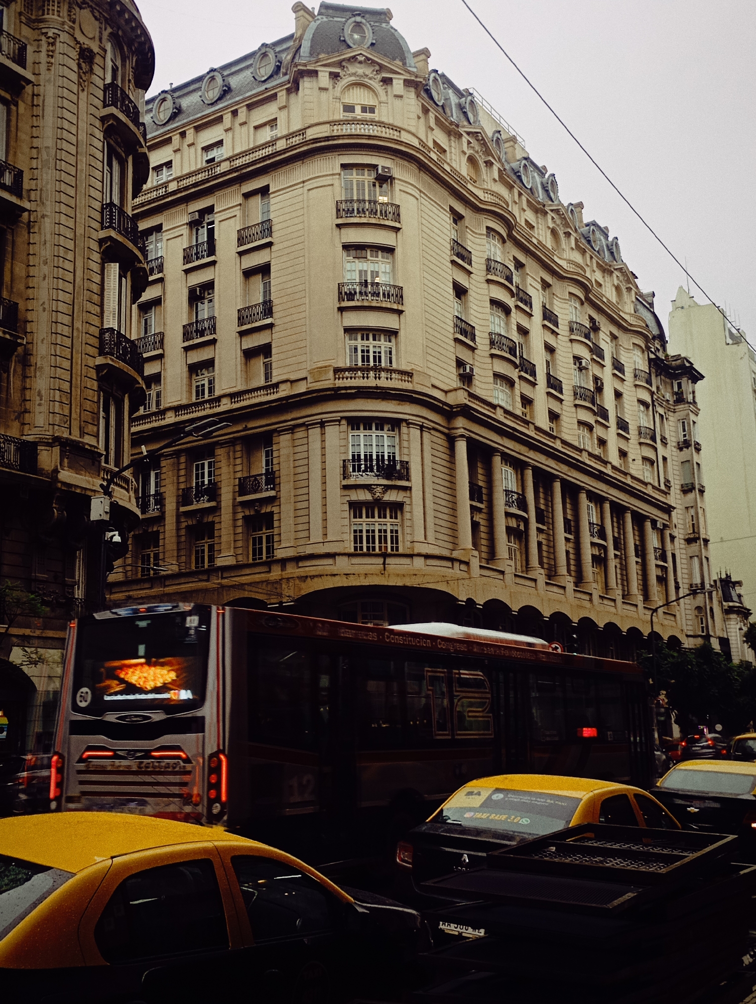 The image presents a bustling street scene in Buenos Aires, Argentina, characterized by grand architecture and urban traffic under an overcast sky.
The prominent subject is a large, ornate building occupying the left and center of the frame. Its classical, European-influenced design features a light-colored stone facade with multiple stories, rounded corners, and a mansard-style roof adorned with circular dormer windows. Intricate details include balconies with dark iron railings, decorative pilasters, and cornices, with large windows visible on every floor. Another, less ornate building stands to the far left, while a taller, more modern-looking structure rises in the background on the right.
In the foreground and mid-ground, a stream of city traffic is visible. A large, red and silver city bus is central, viewed from the rear, displaying a bright, illuminated orange advertisement, possibly for food like pizza, on its back screen. Below this, partially visible text includes "AMOR" and "COLLAGE." The side of a second bus is also visible, showing route details like "Constitución - Congreso -" and the number "12" for its line. Several iconic yellow-roofed Buenos Aires taxis are positioned in the foreground and among other vehicles. One taxi's rear window clearly displays "TAXI BAJE 2.0" and a WhatsApp logo.
The overall lighting is soft and diffused due to the cloudy weather, giving the scene a muted, somewhat muted appearance, characteristic of an overcast day. No specific individuals are clearly discernible, but the presence of public transport and taxis indicates a lively urban environment. The scene captures the blend of historic European-style architecture with the daily movement of a modern South American metropolis.