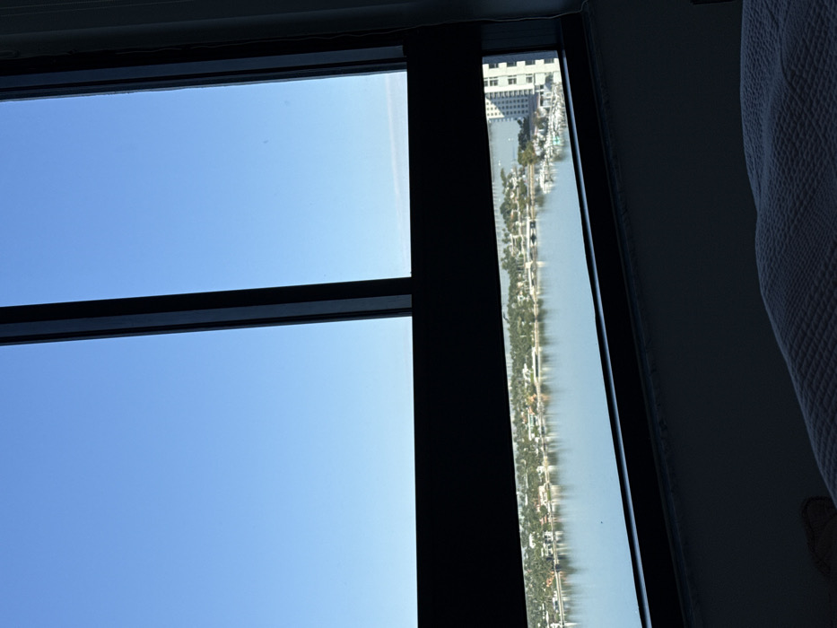 The image displays a high-angle view from an interior room, likely a bedroom, through a large, dark-framed window. The immediate foreground features a light-colored, textured bedspread. The window structure consists of prominent dark vertical and horizontal mullions that segment the exterior view. Beyond the window, a wide expanse of calm, blue water, consistent with a bay or intercoastal waterway, occupies the mid-ground. The water reflects a clear, bright blue sky, which dominates the upper portion of the frame. Across the water, a verdant shoreline is visible, featuring numerous low-rise residential structures with light-colored roofs nestled among dense tropical vegetation. To the right, a marina contains multiple white boats docked, and a multi-story white building is partially observed. A bridge or causeway is visible crossing the water further in the distance. No individuals are present. The lighting suggests daytime. This landscape configuration is characteristic of Miami Beach, Florida, United States of America, specifically overlooking Biscayne Bay.