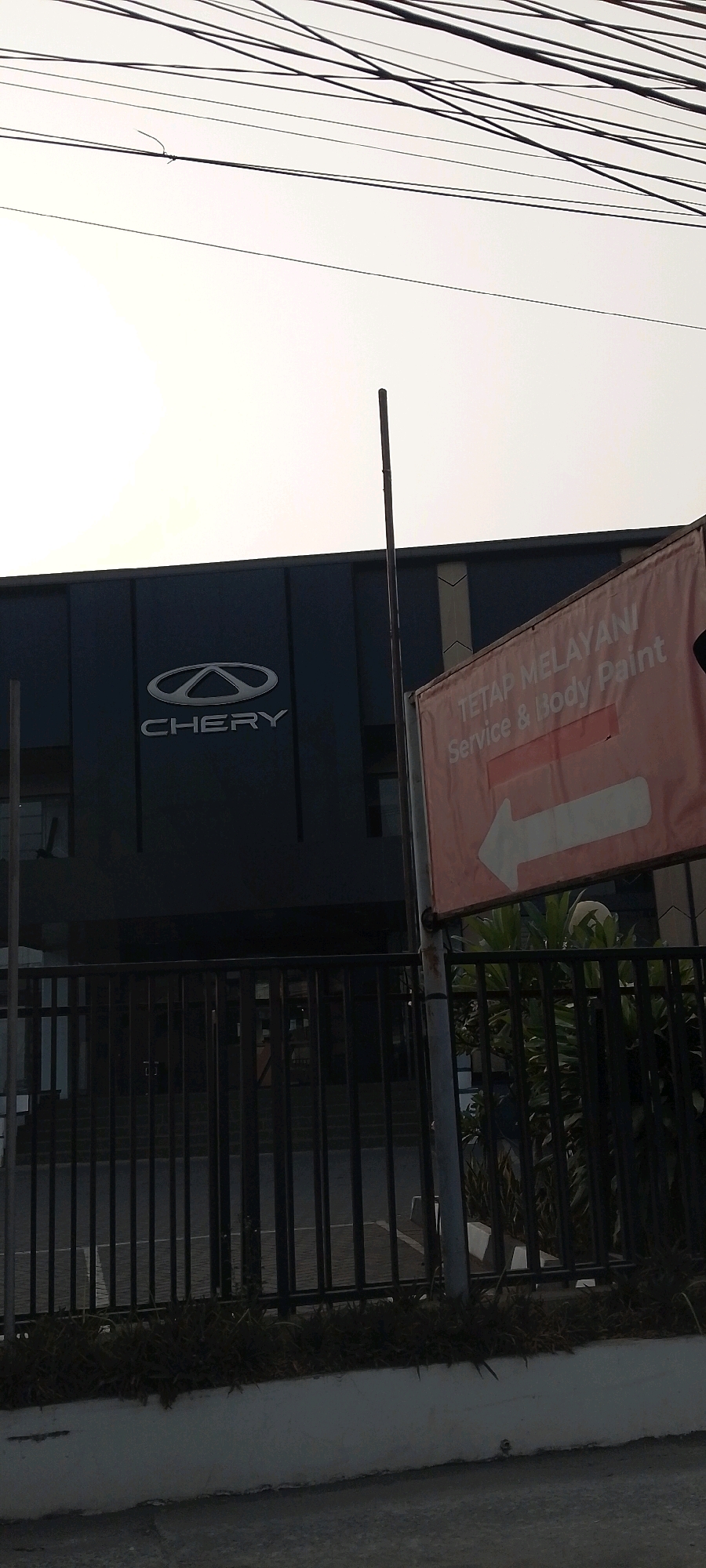 The image shows a Chery dealership in Bekasi, Indonesia. The building has a dark facade with the Chery logo prominently displayed. A red banner with white text on the right side of the image reads "TETAP MELAYANI Service & Body Paint" and features a white arrow pointing to the left. A black metal fence runs across the foreground, separating the street from the dealership's property, which also features some greenery. Overhead, a network of power lines crisscrosses the sky, partially obscuring a bright, overcast sky. There are no people visible in the frame.