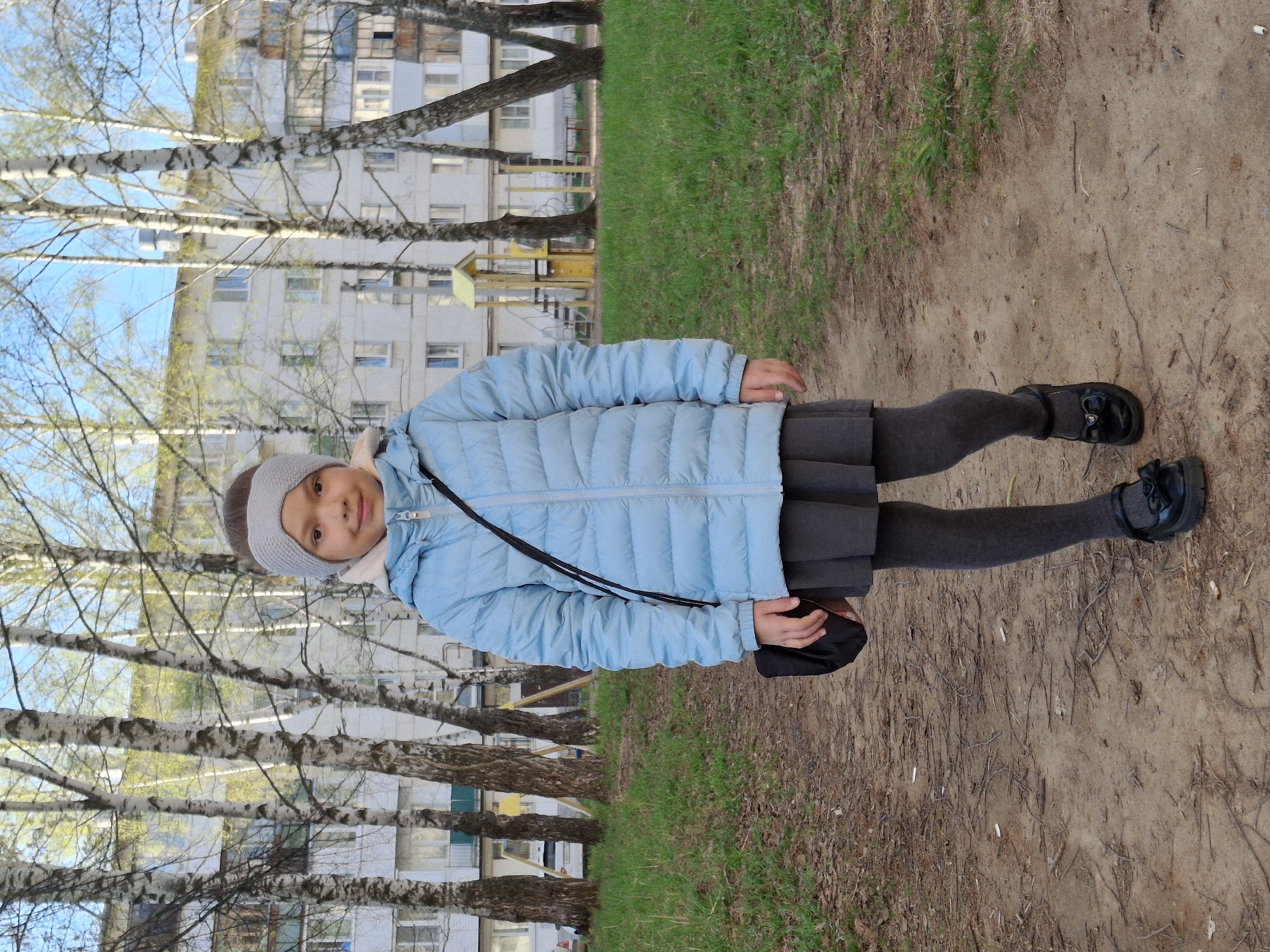 A young girl stands on a dirt path in Chaikovsky, Russia. She wears a light blue puffer jacket, a grey skirt, grey tights, and black shoes. A grey knitted headband adorns her head. She carries a black bag over her shoulder. Behind her are birch trees, grass, and a multi-story building.