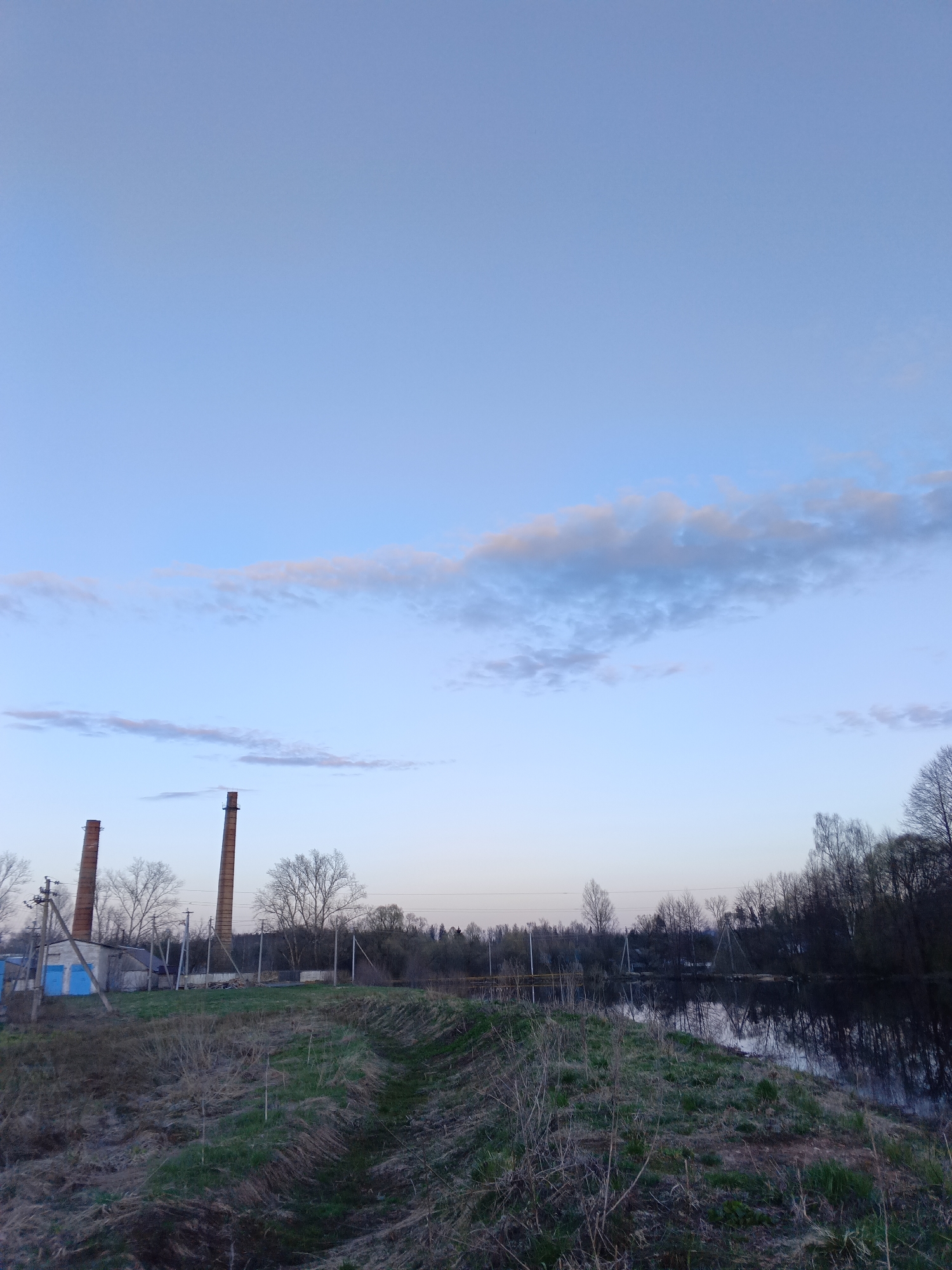 The image shows a landscape near Красный Маяк, Russia. Two tall brick chimneys stand to the left of the frame, adjacent to a small, light-colored building with blue doors. Bare trees surround the buildings and stretch towards the horizon. A dirt path leads from the foreground towards a river or body of water, reflecting the trees on the opposite bank. The sky above is a pale blue with some thin, scattered clouds. The foreground consists of vegetation, a mix of brown dry grass and some green patches.