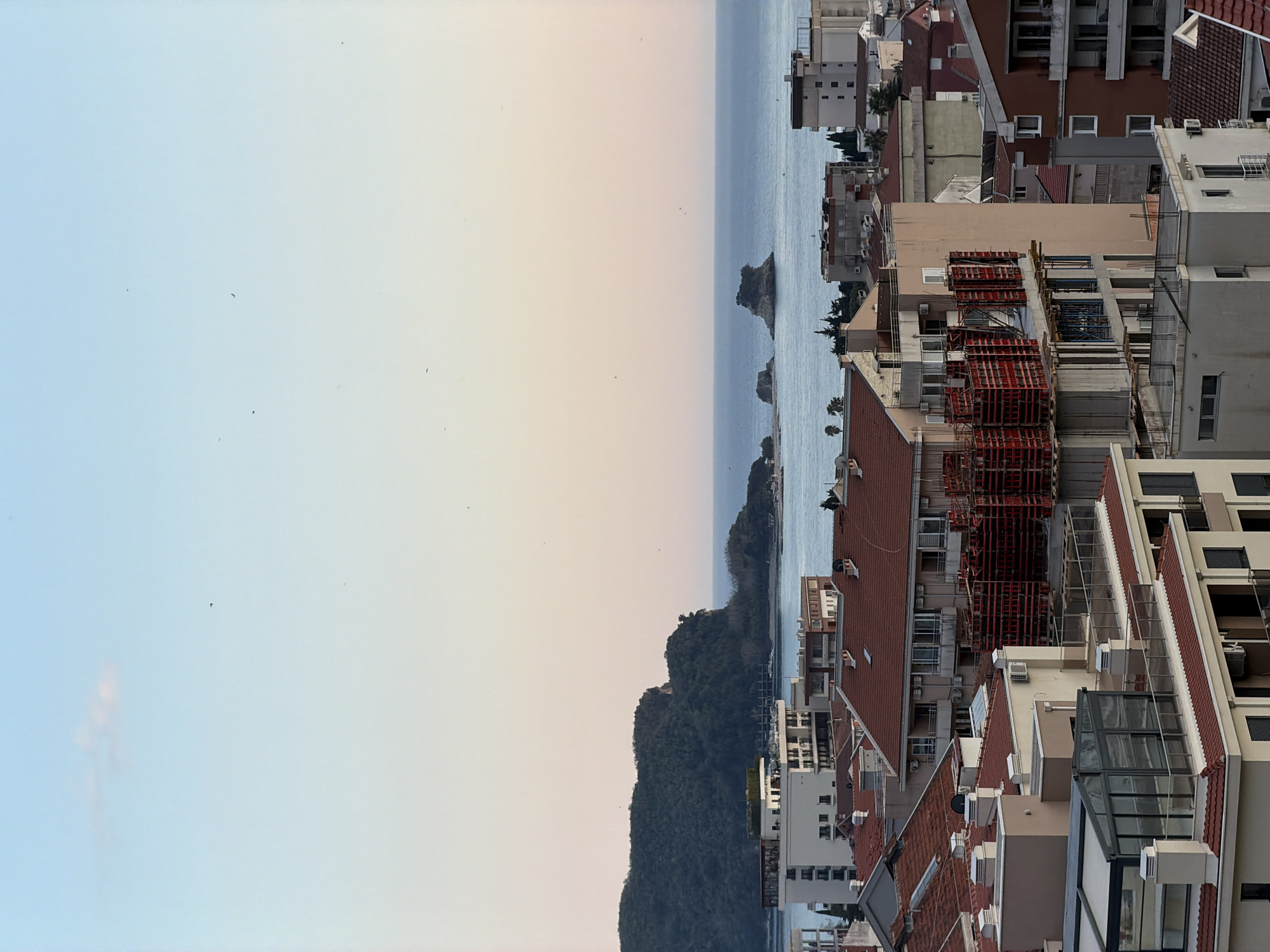 An expansive, high-angle view captures a bustling coastal town, characteristic of Budva, Montenegro, stretching from dense urban development to the serene Adriatic Sea. The image is presented as if rotated to a horizontal landscape orientation, with the sea forming the horizon.
In the foreground and mid-ground, a myriad of multi-story buildings with traditional reddish-brown tiled roofs and facades in shades of white, beige, and brown dominate the scene. Several structures are clearly under active construction, featuring prominent red scaffolding and exposed concrete elements, indicating ongoing development in the area. Details such as air conditioning units, balconies, and windows are visible on the building exteriors.
The tranquil, blue-grey sea occupies the middle distance, extending to a slightly hazy horizon. Scattered across the water are several small, rocky islets. To the left, a large, dark green, forested mountain or hill rises dramatically from the coastline.
The sky above is clear and vast, displaying a soft gradient from light blue on the left to a warm, peachy hue on the right, suggesting the gentle light of either early morning or late afternoon. A few tiny, dark specks, possibly distant birds, are visible against the expansive sky.
No discernible human figures or specific activities are captured, aside from the implied construction work. No legible text or signage is apparent on any of the visible buildings. The overall impression is one of a vibrant coastal settlement blending urban expansion with natural beauty.