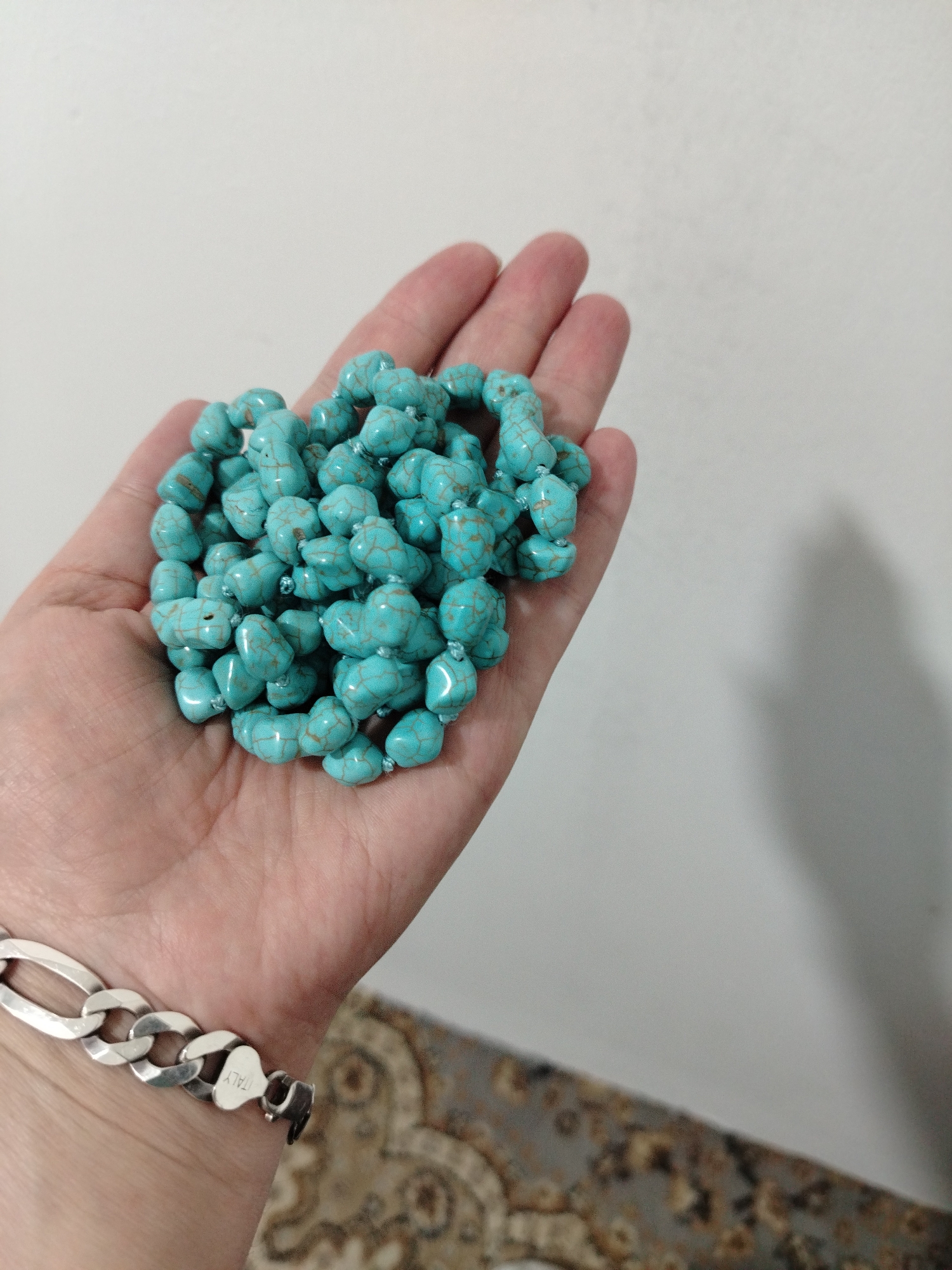 A fair-skinned hand holds a collection of turquoise-colored beads with a brown matrix. The beads appear to be strung together. A silver chain bracelet is visible on the wrist. The background is a white wall with a portion of patterned carpeting visible in the lower right.