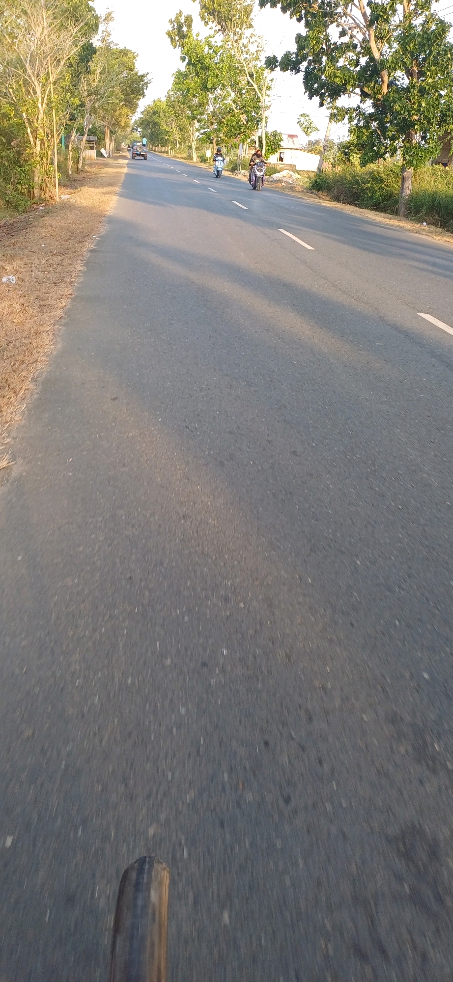 The image depicts a paved road lined with trees and vegetation, likely in Kersik Putih, Indonesia. The road is gray asphalt with white dashed lines marking lanes. In the distance, two motorcycles are visible traveling in the same direction. The motorcycle on the right is ridden by a person wearing a helmet and a dark top. The motorcycle on the left is ridden by a person wearing a helmet and a lighter-colored top. A four-wheel vehicle is also seen further down the road. The sun casts long shadows across the road, indicating it is either early morning or late afternoon. The area to the left of the road is covered in dry grass and sparse trees, while the right side has more dense foliage and a building with a corrugated roof. The foreground of the image shows the tire and part of the frame of what appears to be a bicycle, suggesting the photo was taken from a moving bicycle.