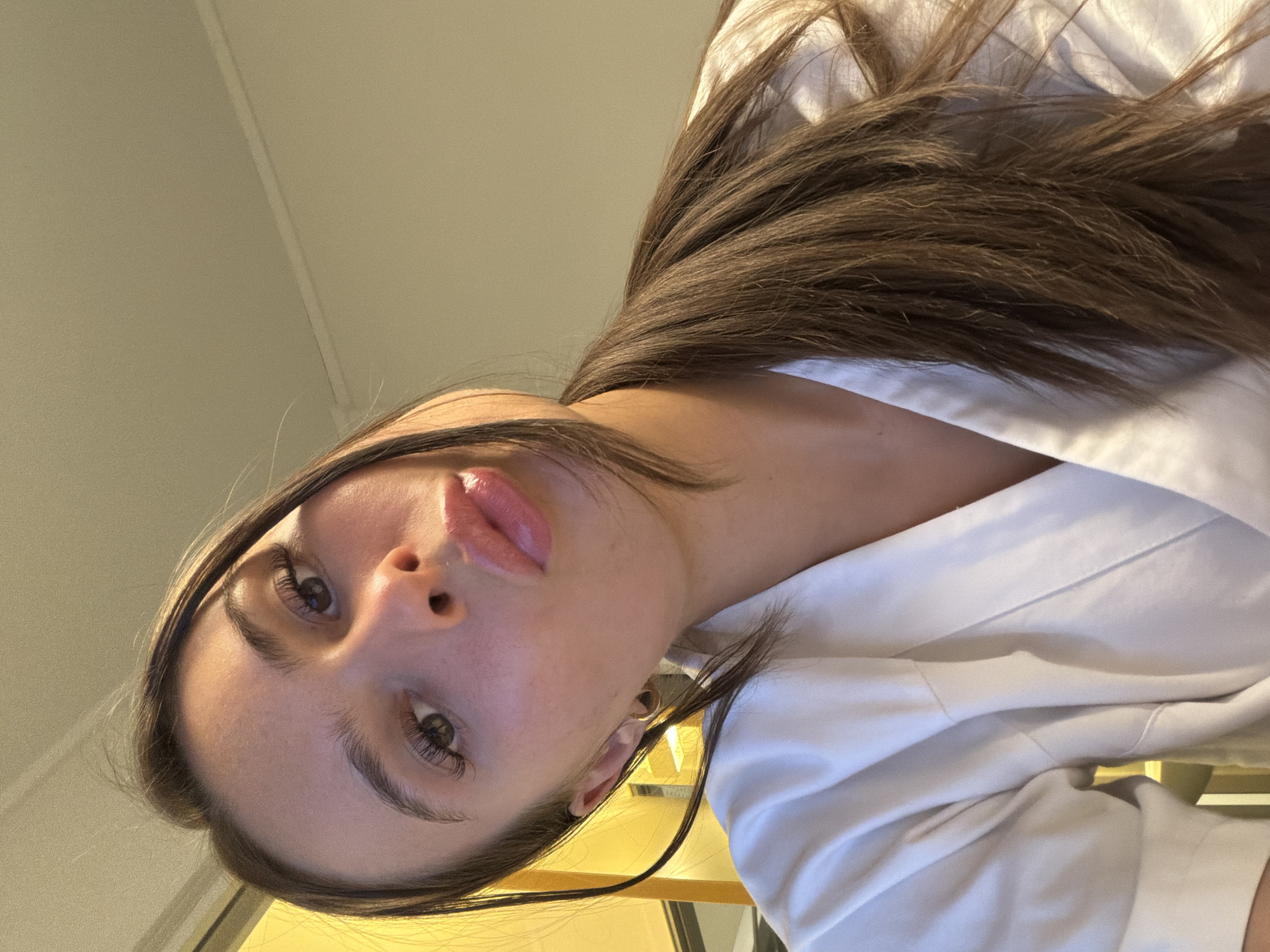 A young woman with long, dark brown hair, tied in a ponytail, poses for a selfie. She is wearing a white robe. Her face is angled towards the camera, and her lips are pursed. She has on what appears to be light makeup, including mascara and lip gloss. Her expression is playful. The background is blurred and appears to be an indoor setting with warm lighting, possibly a hotel room. The image is cropped to focus on her face and upper body. The image does not contain any information to indicate it is located in Harstad, Norway.