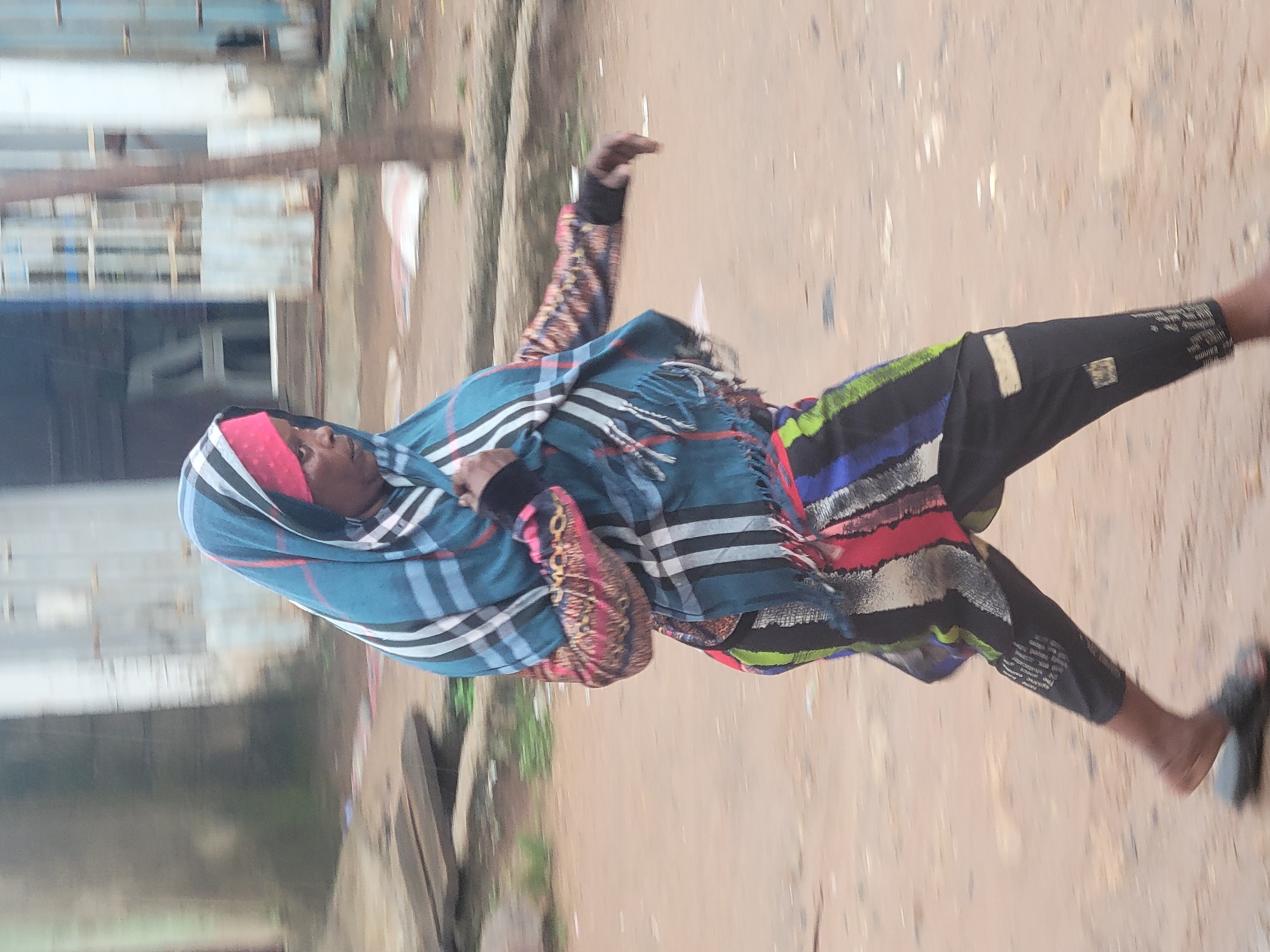 An adult woman in motion is captured in a medium shot, walking outdoors on a dirt path in Madalla, Nigeria. She is wearing a pink head covering, a blue and black plaid scarf draped over her shoulders, a long-sleeved shirt with colorful patterns on the sleeves, and black trousers with vertical colorful stripes. Her left arm is bent, and her hand is extended forward. The background shows a blurred environment with buildings and trees. The image has motion blur, indicating movement.