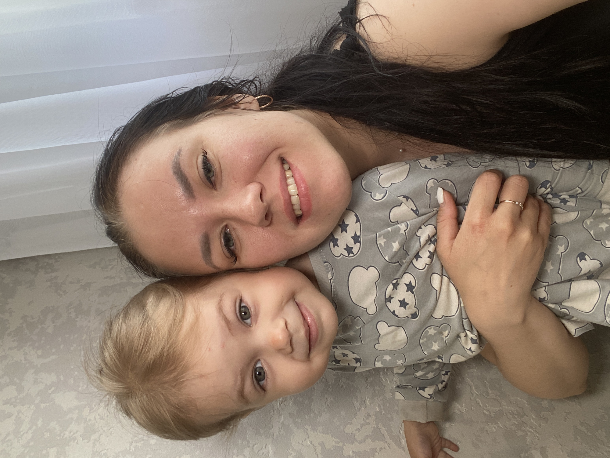 A selfie shows a woman with dark, long hair and a fair-skinned child with blonde hair, in an embrace. The woman is smiling, and the child is looking at the camera with a small smile. The child is wearing a grey patterned pajama top, and the woman has a gold ring on her finger. The background includes a white curtain and a grey wall. There are no indications of the city of Chaikovsky, Russia.