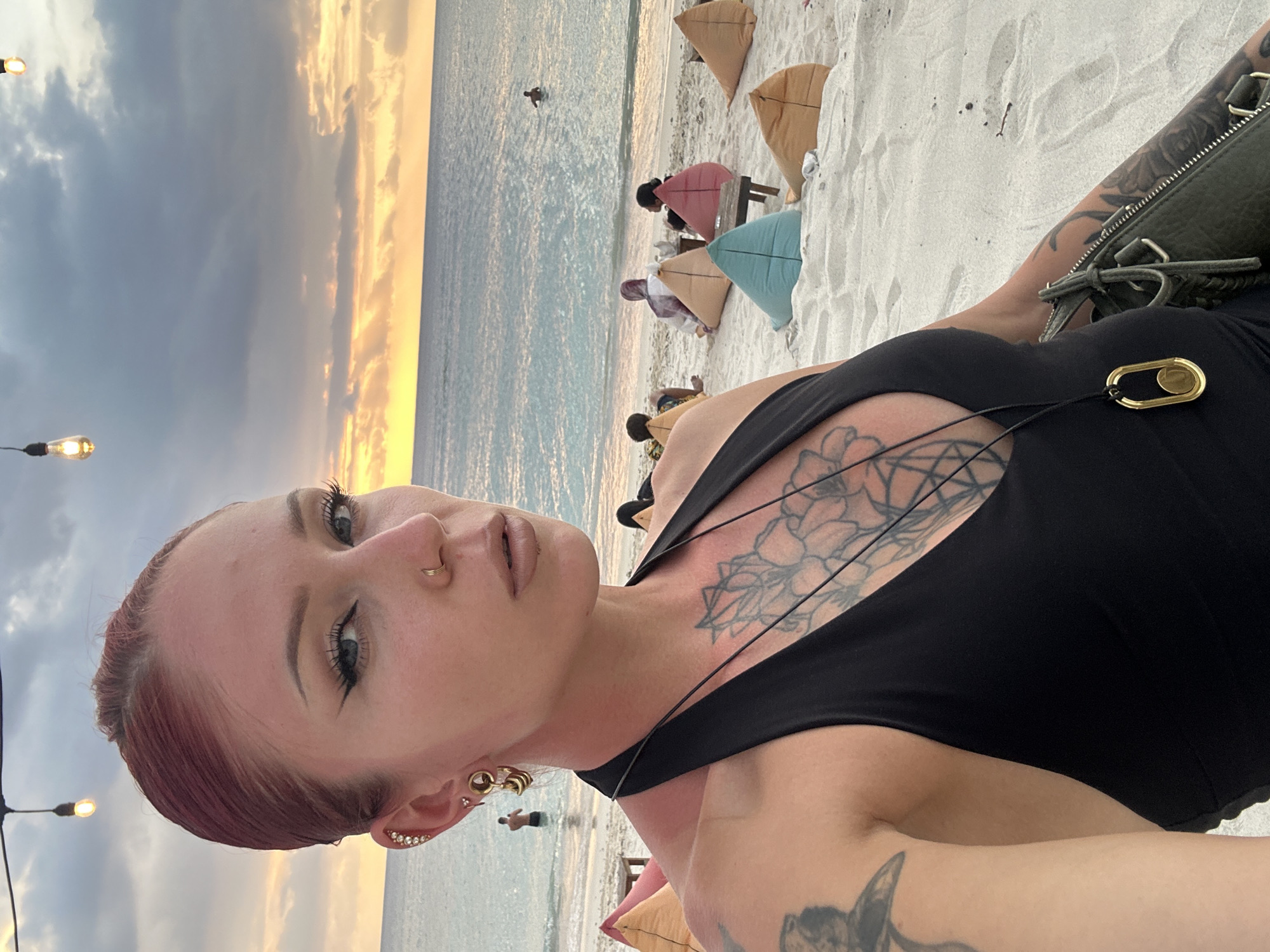 The image captures a scene on a beach in an unknown location in Indonesia, at what appears to be sunset. A light-skinned person is taking a selfie. They have visible tattoos on their chest and arm, short red hair, and several piercings on their ear and nose. They are wearing a black top. In the background, people are relaxing on beanbag chairs. The sea and the sky at sunset are visible behind them. Hanging light bulbs add a decorative touch to the scene.