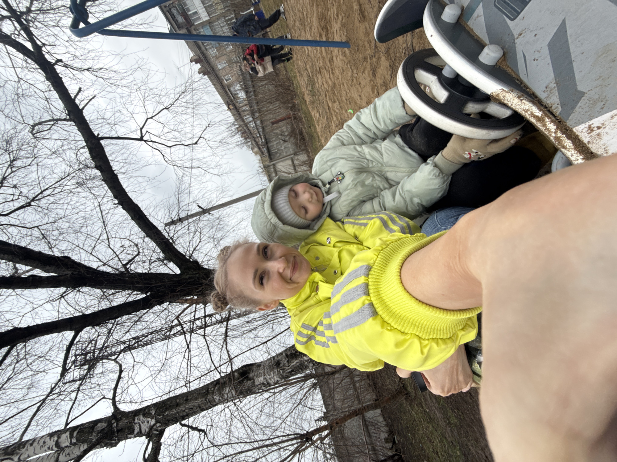 A woman in a yellow jacket and a child in a light green jacket are on a playground. The woman has her hair in buns. The child is wearing a hooded jacket with a grey knit hat underneath and gloves. They are sitting in a vehicle-shaped play structure with two black and white steering wheels. Behind the structure, other children are on playground equipment. A building and bare trees are in the background. The location is in the city of Perm, Russia.