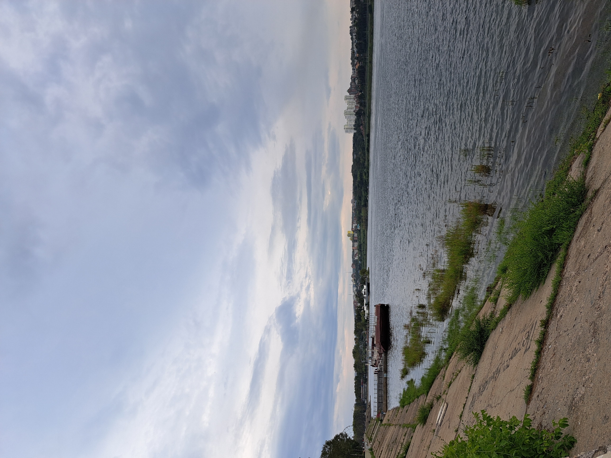 The image depicts a wide, calm river flowing through a city under a cloudy sky. On the left bank, a sloped concrete embankment is visible, with patches of grass growing along its edge. Several moored boats and a pier are situated along the embankment. In the background, a green treeline separates the river from a cityscape featuring a cluster of multi-story buildings and other structures. The water is dark and reflects the muted light of the overcast sky. The scene appears to be a waterfront area in Irkutsk, Russia.