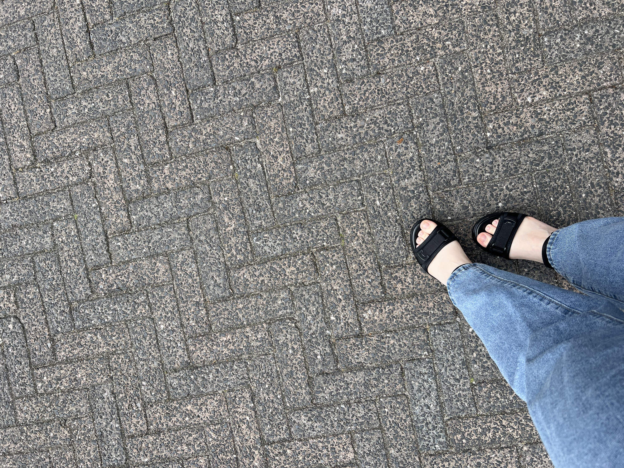 The image shows a person wearing blue jeans and black sandals standing on a herringbone-patterned paved surface. The surface appears to be made of rectangular grey pavers with a rough texture. The person's feet are visible in the lower half of the frame, with their left leg extending upwards and to the left, and their right leg extending slightly upwards and to the right. The lighting suggests it is daytime. The location is identified as Grou, Netherlands, though there are no specific landmarks or clues in the image to confirm this beyond the provided information.