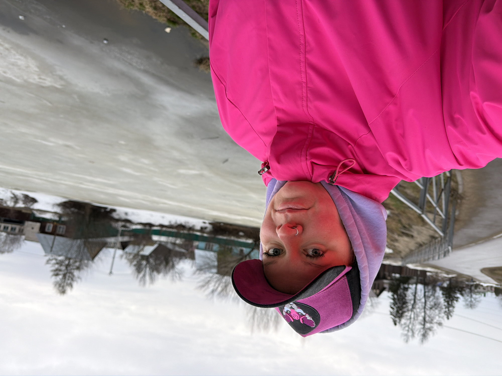 A woman in a pink jacket and cap stands outside. She is facing the camera. Behind her is a frozen lake or body of water. On the far side of the water, there are residential buildings, consistent with an urban area. The scene is consistent with the city of Chaikovsky, Russia. A fence and a road are also visible.