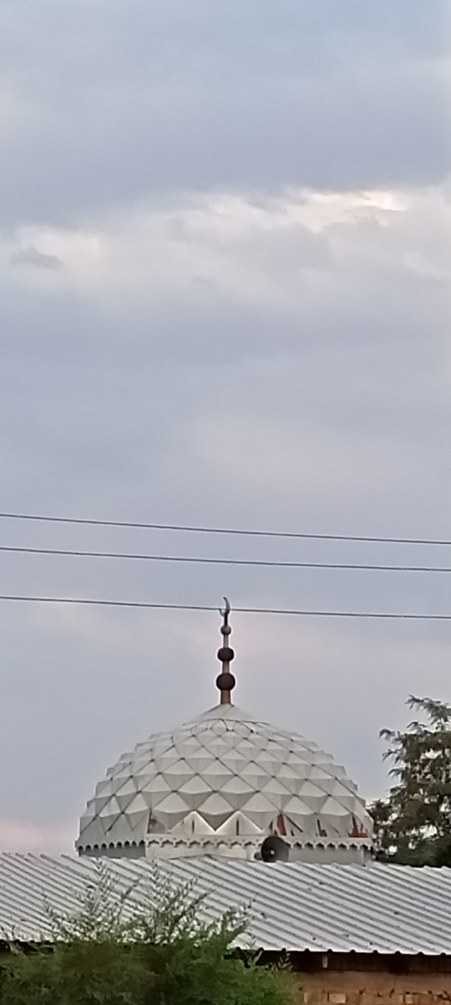 The image shows the top of a mosque dome with a crescent moon finial, located in Sokuluk, Kyrgyzstan. The dome is white and has a geometric pattern. Above the dome, power lines stretch horizontally across the frame. The sky is overcast with clouds. Below the dome, the metal roof of the mosque is visible, with some greenery in the foreground. There are no people visible in the image.