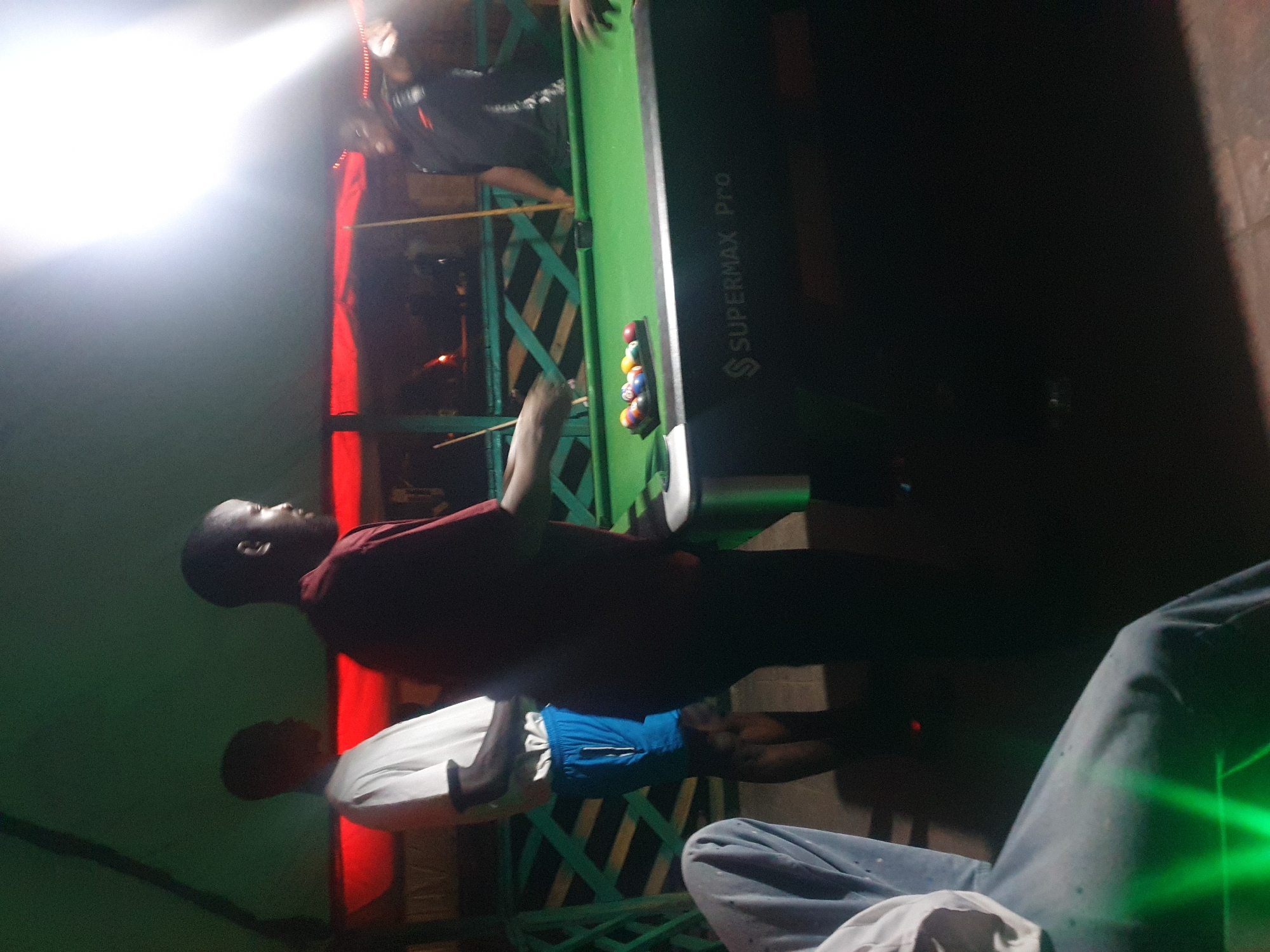 In Lokoja, Nigeria, a dimly lit indoor setting features a group of men playing pool. A man in a red shirt, facing away from the camera, stands to the right of a pool table, cue in hand, as if about to take a shot. To his left, another man in a white t-shirt and blue shorts, also facing away, observes the game. The pool table, green felt, has a rack of colorful balls positioned on it. The background shows a red canopy overhead and green lattice fencing. The lighting is low, with a bright light source to the upper right, casting a glare and creating shadows.