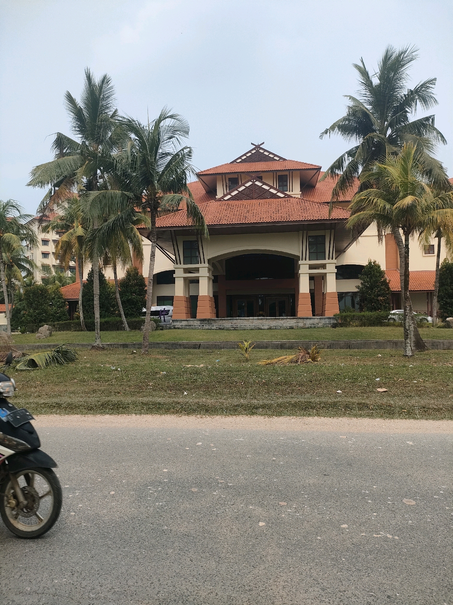 The image depicts a tropical resort building with a red tiled roof, reminiscent of Balinese architecture, situated in Batam, Indonesia. The building features a grand entrance with columns and a wide arched opening leading into the interior. Tall palm trees with lush green fronds are strategically planted around the property, framing the structure and adding to the tropical ambiance. In the foreground, a motorcycle is partially visible, suggesting it is either parked or passing by on the adjacent road. The surrounding environment is characterized by a manicured lawn, some scattered fallen palm fronds, and a hazy sky. The overall scene conveys a sense of a holiday destination, common in Batam's resort areas.