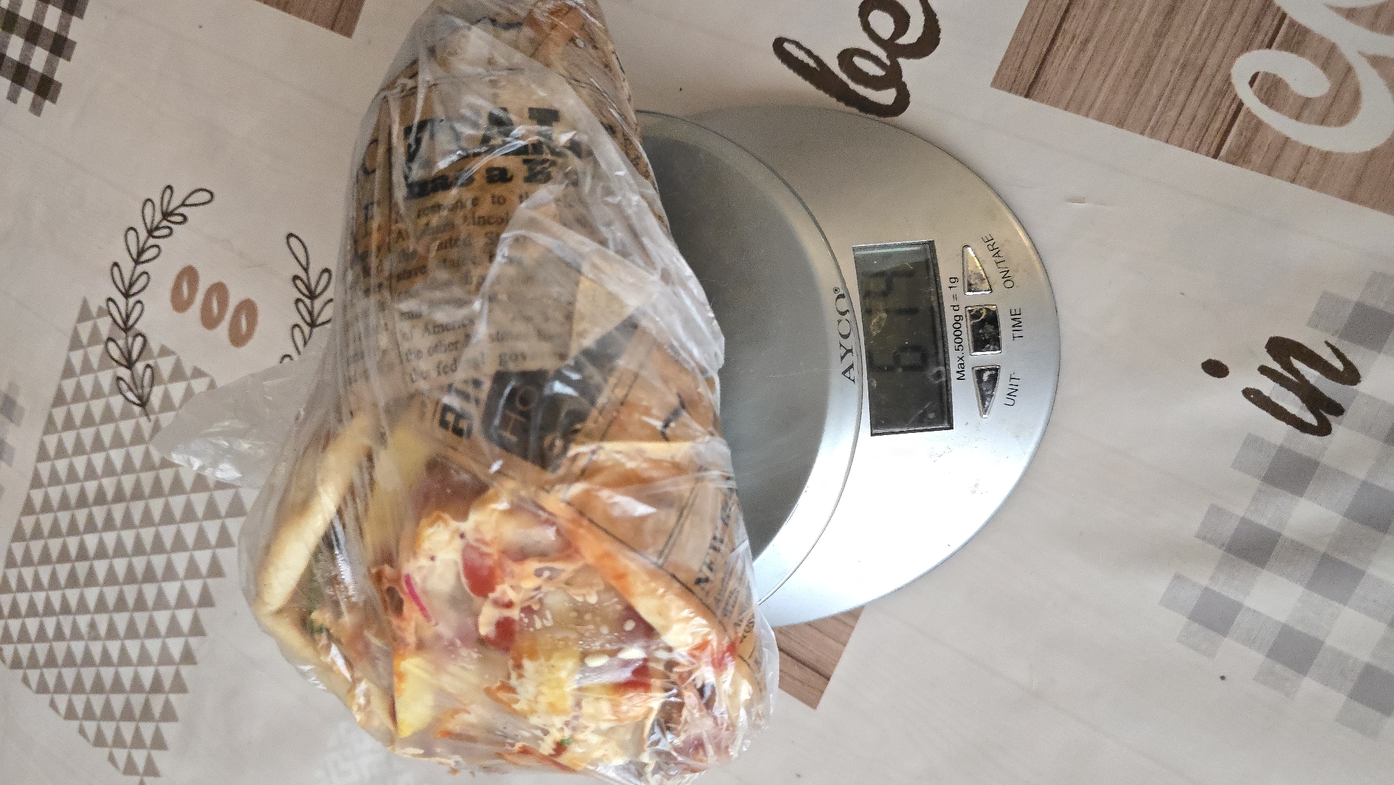 A food item, possibly a wrap or burrito, is encased in clear plastic and placed on a digital kitchen scale. The scale displays a weight of 674 grams. The scale itself is silver and oval-shaped, with buttons labeled "UNIT", "TIME", and "ON/TARE". The food item appears to contain various ingredients, including what looks like meat, vegetables, and sauce. The scale is situated on a table with a patterned tablecloth. The tablecloth features a geometric design in shades of beige and grey, along with some decorative floral and circular motifs. In the background, the pattern on the tablecloth transitions to a wood-grain texture. The overall lighting suggests an indoor setting.