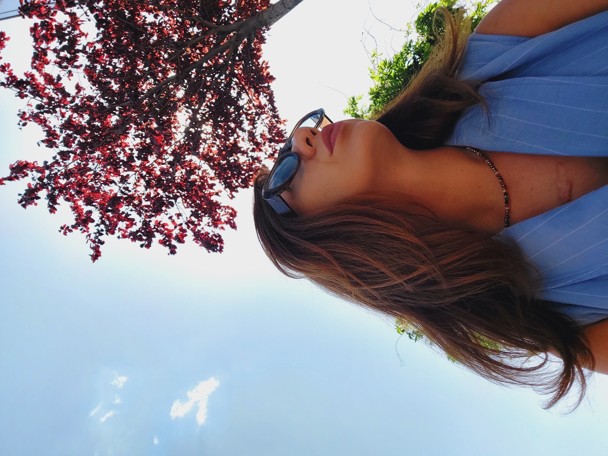 This is a low-angle, close-up, eye-level shot of a woman wearing sunglasses and a blue pinstriped vest, looking up towards a tree with dark red leaves against a bright blue sky with scattered white clouds. The woman has long, brown, wavy hair and is wearing a beaded necklace. A scar is visible on her chest. The scene appears to be outdoors, possibly in a park or garden, in Sofia, Bulgaria.