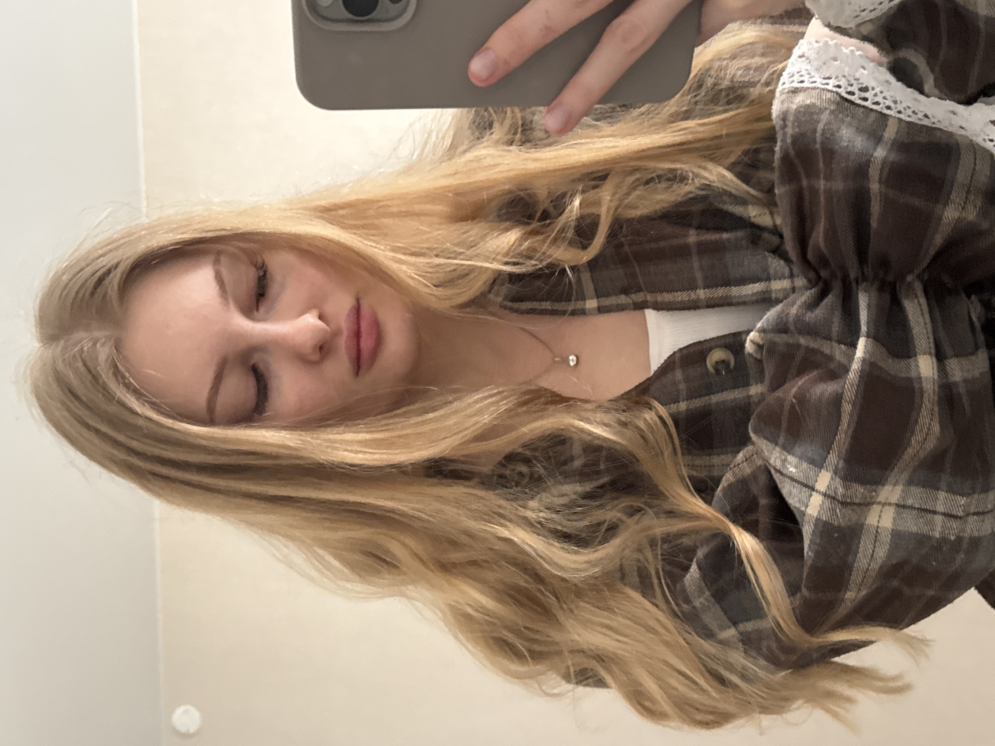 A young woman with long, blonde hair takes a selfie indoors using a grey smartphone. She wears a brown and beige plaid shirt with lace trim at the sleeve. A silver necklace with a small pendant is visible. The background appears to be a plain, light-colored wall. It is impossible to determine the location in Perm, Russia from the provided image.