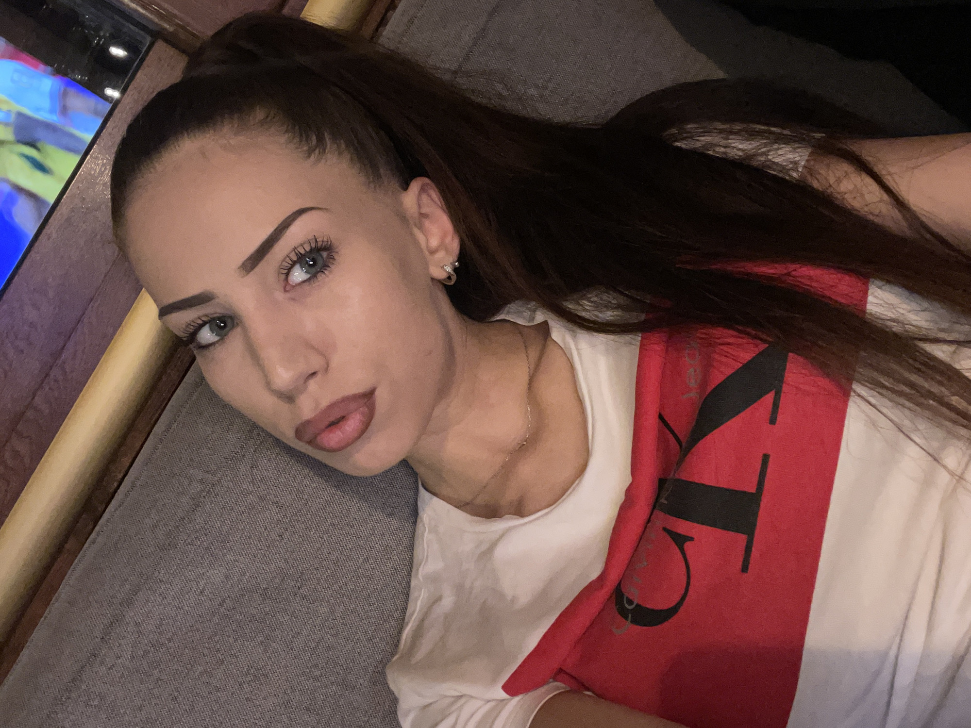 A woman with long, dark brown hair pulled into a high ponytail is lying down, resting her head on a grey textured surface, likely a couch or bed. She is wearing a white t-shirt with a red stripe across the chest featuring large black lettering. The text visible on the stripe appears to be part of a brand logo. She has light-colored eyes, dark eyebrows, and is wearing a silver hoop earring in her visible ear. A thin silver necklace is visible around her neck. The lighting suggests an indoor setting, possibly in the evening or with artificial light. The overall mood of the image is relaxed and casual. To the left of the woman, a portion of a screen displaying colorful images is visible, suggesting she might be near a television or computer.