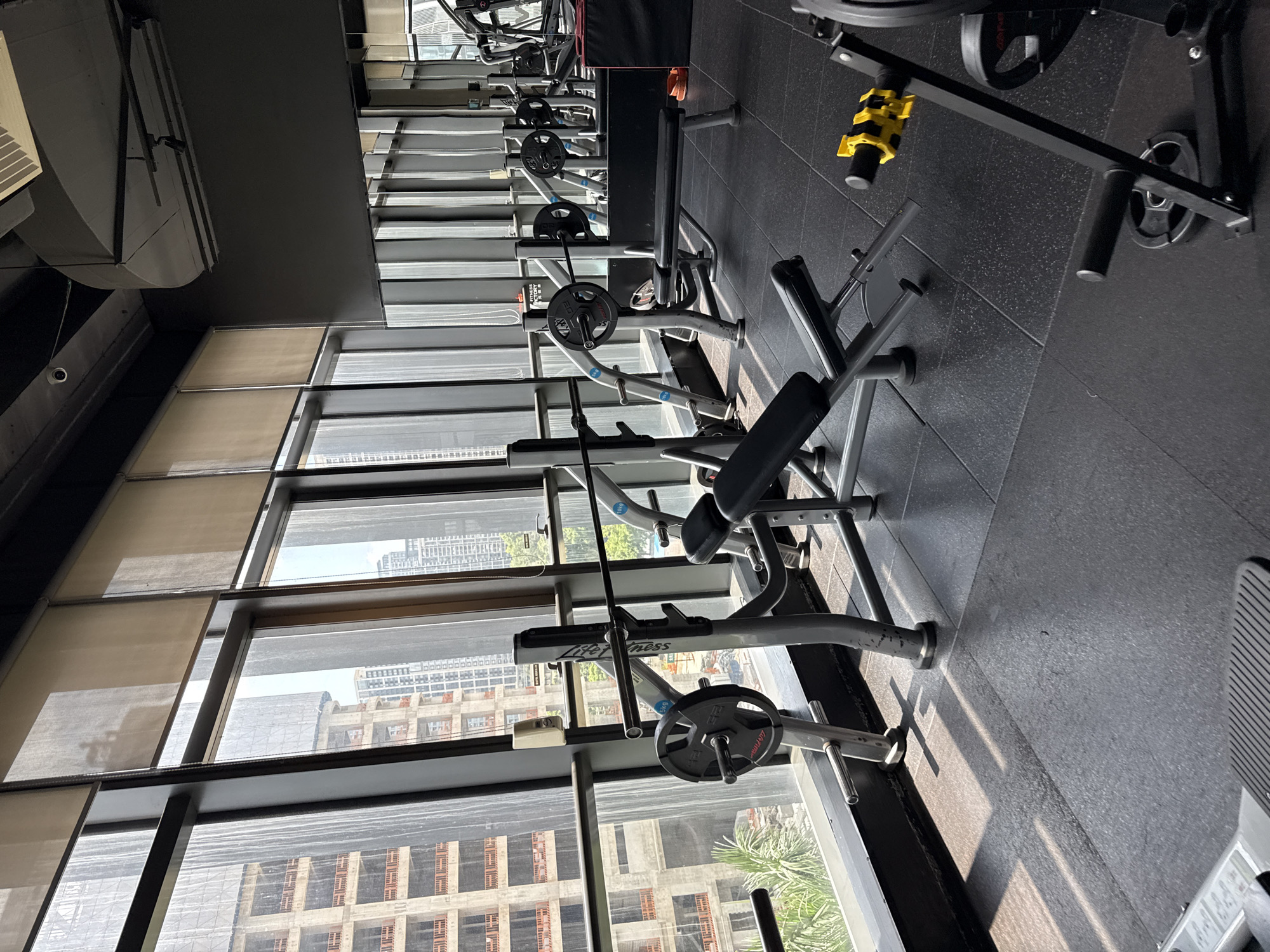 The image shows an indoor gym with large windows overlooking a cityscape. The gym is equipped with various weightlifting machines, including bench presses with barbells and weight plates, and adjustable benches. The floor is made of black rubber tiles. Sunlight streams through the windows, casting shadows on the floor. In the background, several tall buildings are under construction, suggesting an urban environment in a developing area. The overall atmosphere is that of a modern, well-equipped fitness facility. The location is identified as Haizhu District, China. There are no people visible in the image.