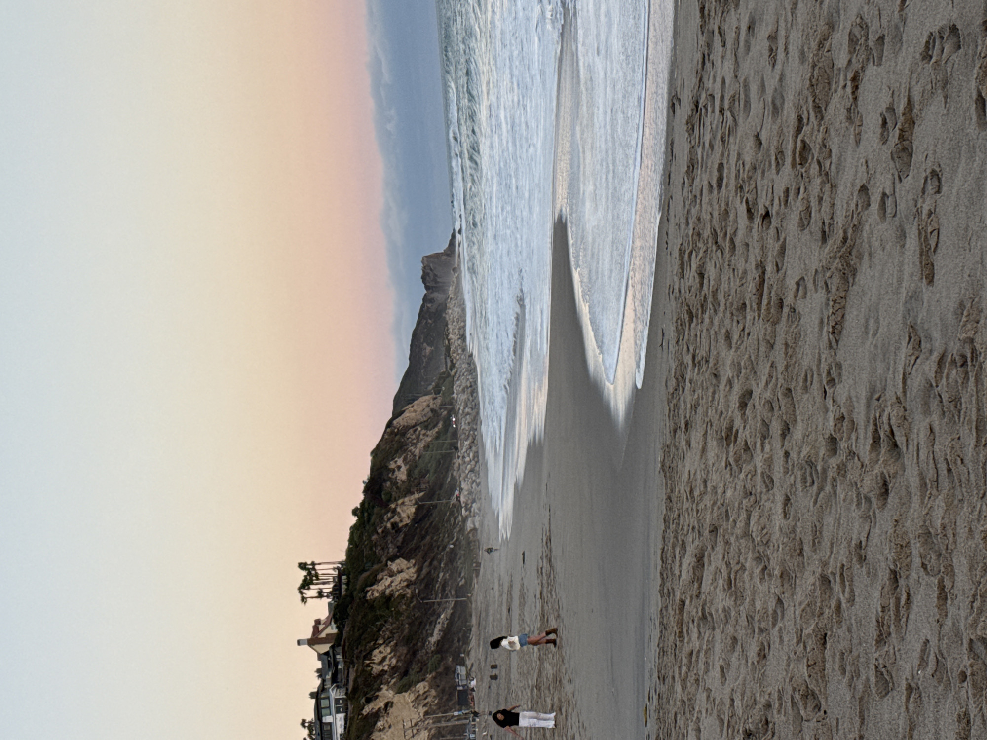 The image depicts a beach scene in Malibu, United States of America, during what appears to be sunset or dawn, given the soft gradient of colors in the sky, transitioning from pale blue to pink and orange. In the foreground, the sandy beach is covered in footprints. Further back, two individuals, one wearing white pants and a black top, and another in shorts and a white top, are standing on the sand. A person is also visible riding a bicycle in the middle ground. The middle ground also shows the ocean with waves breaking and a rocky shoreline. In the background, a cliff face is topped with several houses and palm trees, characteristic of the Malibu coastline. The overall atmosphere is serene and peaceful.