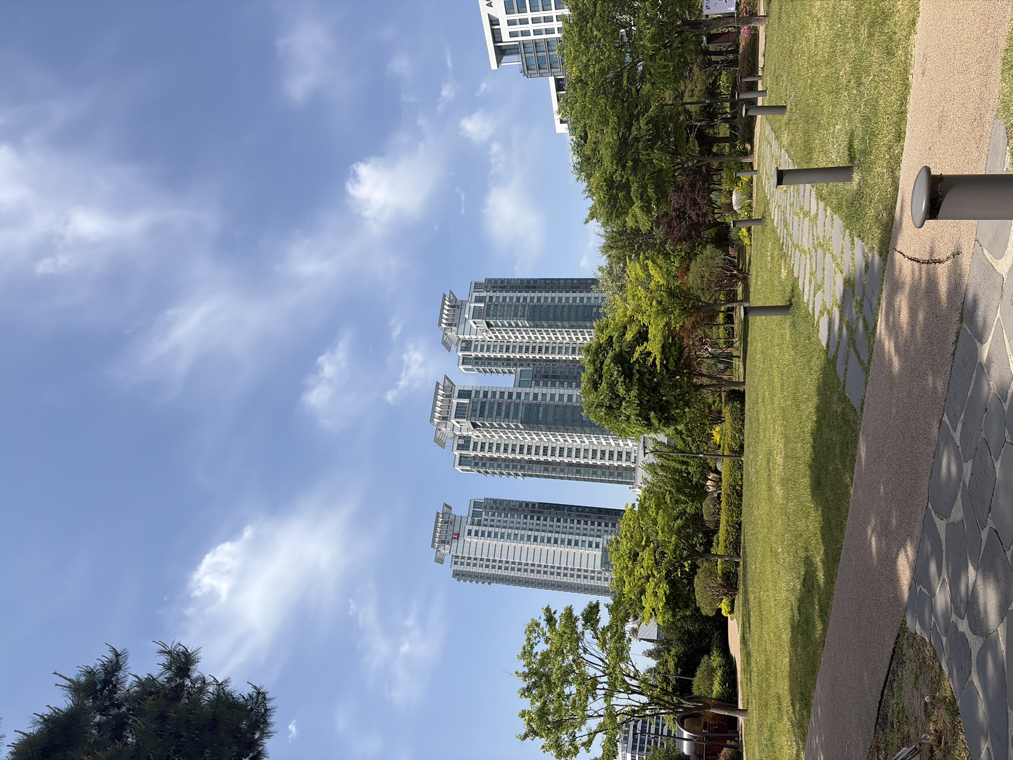 The image shows a park setting in what appears to be a modern area of Seoul, South Korea. Several tall, modern residential buildings are visible in the background, indicating an urban setting. The park has well-maintained grassy areas, trees, and a paved walkway with embedded stone tiles, and evenly spaced lamp posts line the pathway. The sky is blue with scattered clouds. There are no people visible in the image.