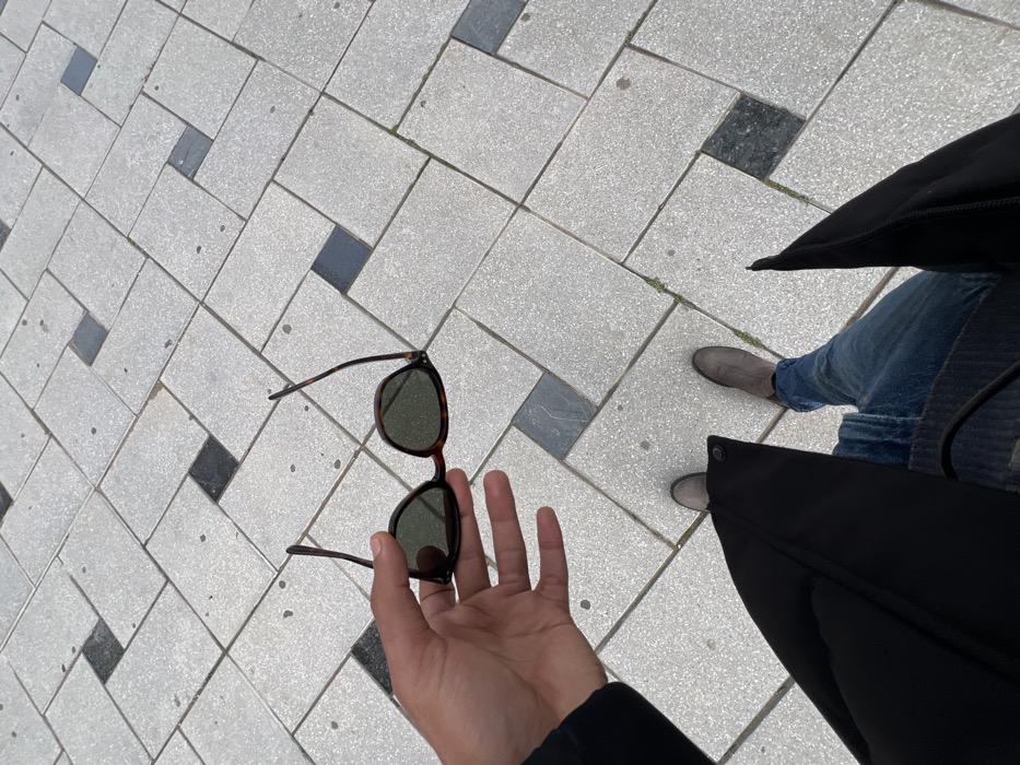 The image depicts an overhead view of a paved outdoor surface in the city of Tanger, Morocco. An individual is visible from the waist down, wearing blue jeans, grey ankle boots, and a black coat or jacket. The person's left hand is extended forward, holding a pair of tortoise-shell framed sunglasses with dark green lenses. The sunglasses' arms are partially open. The ground is paved with light grey rectangular tiles arranged in a repeating pattern, interspersed with darker grey square accent tiles. Minimal organic growth is present in the grout lines between some tiles.