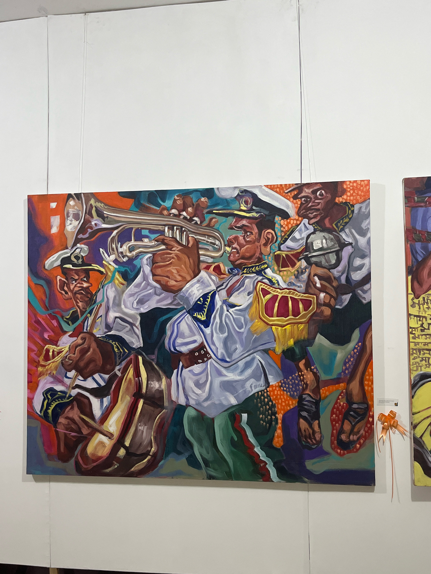 The image shows a vibrant oil painting of a marching band on display in a gallery in Dhaka, Bangladesh. The painting features several individuals in a stylized, somewhat distorted manner, playing musical instruments. In the foreground, a man in a white uniform with gold trim is playing a trumpet, his face contorted in concentration. To his left, another figure, also in a similar uniform, is shown playing a drum with drumsticks. Behind them, a third figure in a white uniform is holding a small cymbal or bell. The background is a chaotic mix of bright colors and abstract shapes, with a textured orange pattern dominating the upper right portion. The painting is hanging on a white wall, with a clear hanging wire visible at the top. A small orange ribbon is attached to the wall to the right of the painting. Another painting, partially visible on the right, has yellow and black elements and appears to have text.