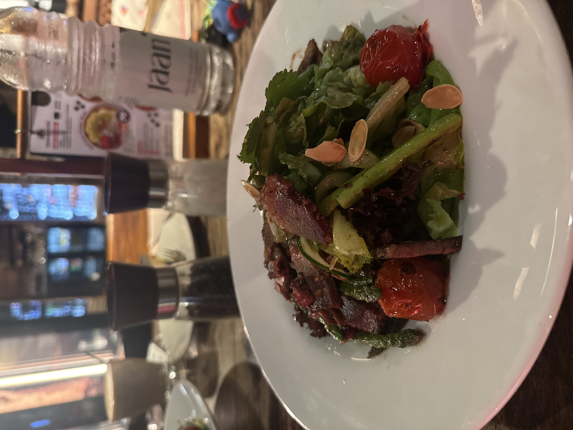 This image, captured in an indoor setting likely a restaurant or cafe in Kuta Selatan, Indonesia, displays a vibrant salad served on a white, oval-shaped plate. The image is rotated 90 degrees clockwise.
The salad features a diverse mix of fresh ingredients. Prominently visible are green leafy vegetables, some darker red leafy greens, and several bright red roasted or grilled cherry tomatoes. Green asparagus spears and pale, oval-shaped sliced almonds are interspersed throughout the salad, adding color and texture. Strips of crispy, dark red-brown bacon are also clearly present among the greens.
To the left of the plate, a clear glass bottle with the word "Jaan" printed vertically on its label is visible. Below this bottle, and partially obscured by it, is a blurred card or menu with some indistinct text and a small food image. Further to the left, two spice grinders, one with a black top and another with a silver top, suggest seasonings available for the meal. The background is softly blurred, indicating a shallow depth of field, with ambient restaurant lighting and reflections creating an intimate dining atmosphere. The scene captures a well-presented dish ready for consumption.