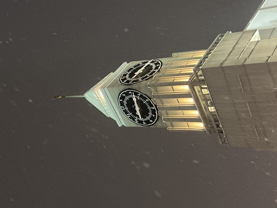 The image captures a modern clock tower at night, standing tall against a dark sky with light snow falling. The photograph is taken from a low angle, looking up at the illuminated structure, and appears to be rotated approximately 90 degrees clockwise.
The tower itself is constructed from light-colored, possibly metallic or stone, panels that give it a sleek, vertical appearance. Several sections of the tower feature horizontal grooves or ribs that are lit from within or by external warm-toned lights, creating a glowing effect that contrasts with the dark surroundings. The top of the tower tapers to a pointed spire, culminating in a slender, dark finial.
Two prominent clock faces are visible on adjacent sides of the tower, both circular with black backgrounds and white markings for the numbers (1 through 12) and minute increments. Both clocks have white minute and hour hands.
Upon closer inspection:
- The clock face positioned lower and to the left (when viewing the image in its original, rotated state, or the left-facing clock if the tower were upright) clearly shows the minute hand pointing directly at the '5', indicating 25 minutes past the hour. The hour hand is just past the '12' mark, indicating the time is approximately 12:25.
- The clock face positioned higher and to the right (when viewing the image in its original, rotated state, or the right-facing clock if the tower were upright) also shows the minute hand pointing directly at the '5', indicating 25 minutes past the hour. However, the hour hand on this clock is just past the '1' mark, suggesting the time is approximately 1:25. This discrepancy in the hour displayed on the two adjacent faces of the same tower is a notable detail.
The sky is a deep, uniform dark gray or black, characteristic of nighttime. Numerous small, diffused white specks are scattered throughout the air, indicative of light snowfall or sleet. The overall scene suggests a cold, quiet winter night.