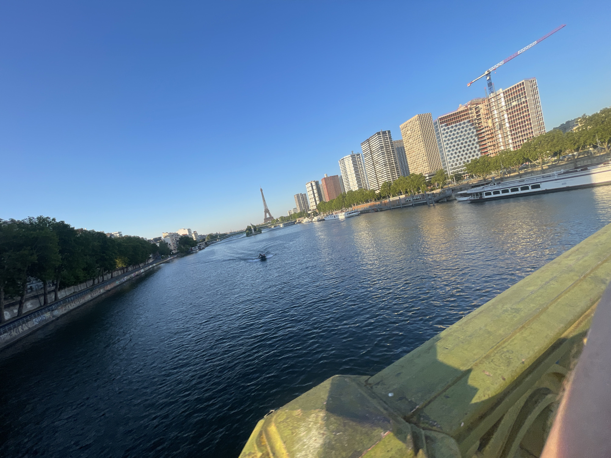 This media file depicts a picturesque view of the Seine River in Paris, France, during what appears to be a clear, sunny day, likely late afternoon or early evening given the quality of light. The scene is captured from the perspective of a bridge, with the foreground showing a section of the bridge's railing, painted a mossy green and showing signs of age.
The river itself is a prominent subject, its surface rippling and reflecting the bright blue sky and surrounding urban landscape. Several boats are visible on the water: a small motorboat with a few occupants can be seen creating a wake further upstream, and larger, white passenger boats are docked or cruising along the opposite bank.
The opposite bank is lined with trees, creating a green buffer between the river and a series of modern buildings. These buildings form a skyline that includes some older, striped high-rises and newer construction projects, one of which is topped by a large red and white construction crane. In the distance, the iconic Eiffel Tower pierces the sky, a clear landmark identifying the location as Paris. The overall mood conveyed is one of peaceful urban life, with the natural element of the river and trees complementing the architectural elements. No visible text is present in the image.