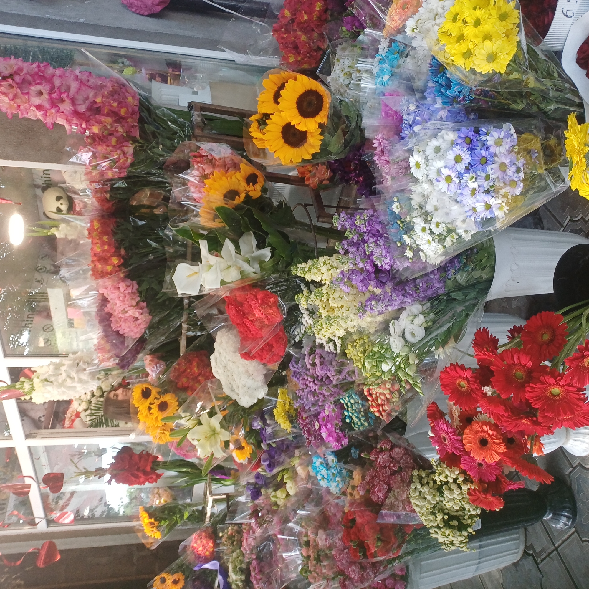 A full shot captures an outdoor flower stand, possibly in a marketplace, displaying a vibrant array of freshly cut flowers. The stand features a wide variety of colorful blooms, including sunflowers, gerbera daisies, lilies, and an assortment of smaller, clustered flowers in shades of pink, purple, yellow, red, and white. Many of the bouquets are wrapped in clear plastic, common for floral arrangements. In the background, a glass-paned window reveals the interior of a shop, with glimpses of shelves and decorative items, including a panda plush toy. A young woman with dark hair is partially visible behind the flowers, looking towards the interior of the shop. The ground is paved with grey tiles. The scene appears to be during daylight hours, with ample natural light illuminating the flowers.