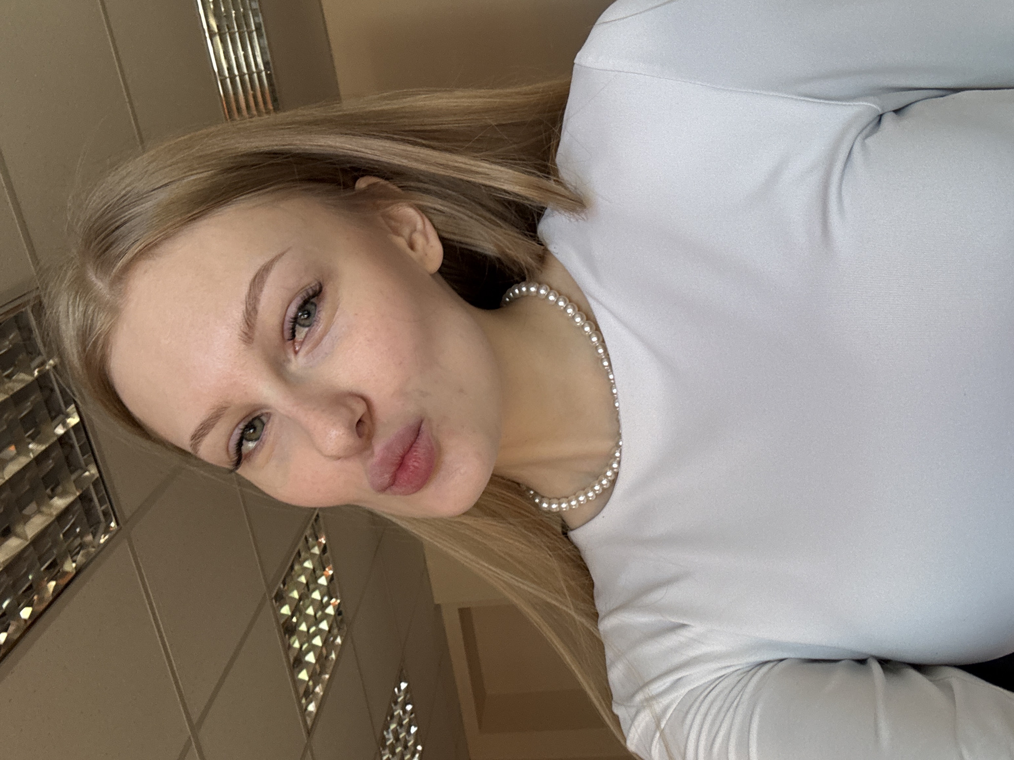This is a diagonal portrait of a young woman with long, straight blonde hair, fair skin, and light green eyes. She is looking directly at the camera with a playful pursed-lip expression, often referred to as a "kiss face." Her eyebrows are neatly shaped, and her eyelashes are visible.
She is wearing a white, long-sleeved top, possibly made of a smooth, stretchable fabric. Around her neck, she has a delicate pearl necklace.
The background indicates an indoor setting, likely an office or a commercial space, characterized by a light-colored ceiling with square tiles. Several recessed lighting fixtures are visible on the ceiling, featuring a grid-like, reflective interior design that creates distinctive patterns of light. The walls appear to be a plain, light brown or beige. The lighting is bright and even.
The activity depicted is a self-portrait or selfie. The overall mood is casual and playful. There is no visible text in the image. The location context for this scene is Perm, Russia.