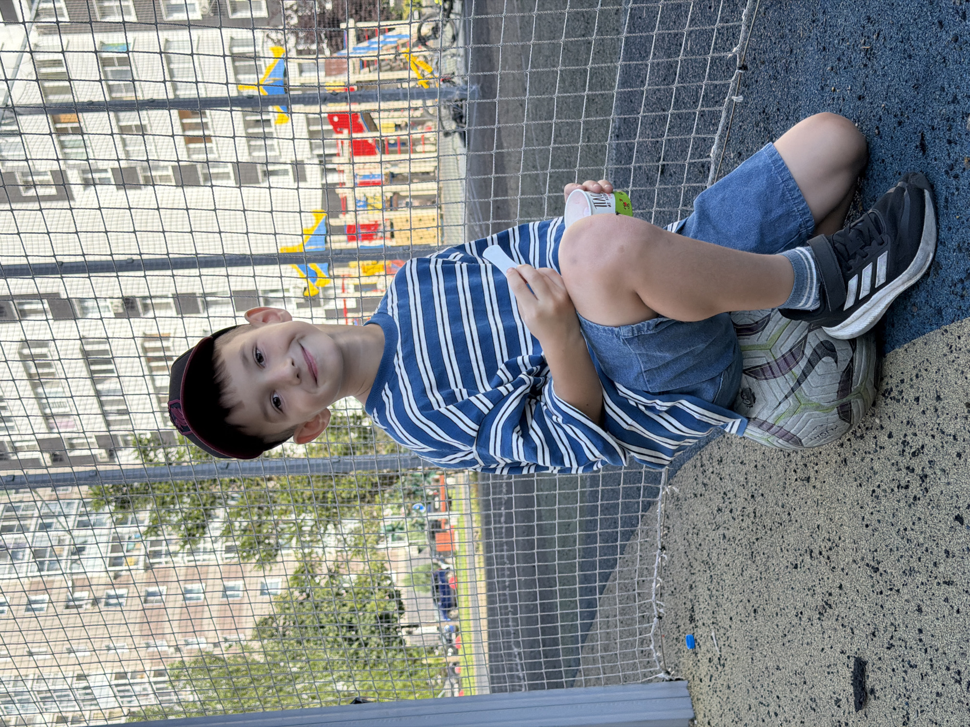 The image shows a young boy wearing a blue and white striped t-shirt, denim shorts, a baseball cap, and black sneakers with white accents. He is sitting on a soccer ball on a playground with a mesh fence in the foreground and buildings in the background. The boy is holding a small white object in his right hand and a cup in his left. The playground appears to be part of a residential area, possibly in Kazan, Russia, given the architecture of the buildings and the presence of a children's playground. The overall setting suggests an outdoor recreational area within a city.