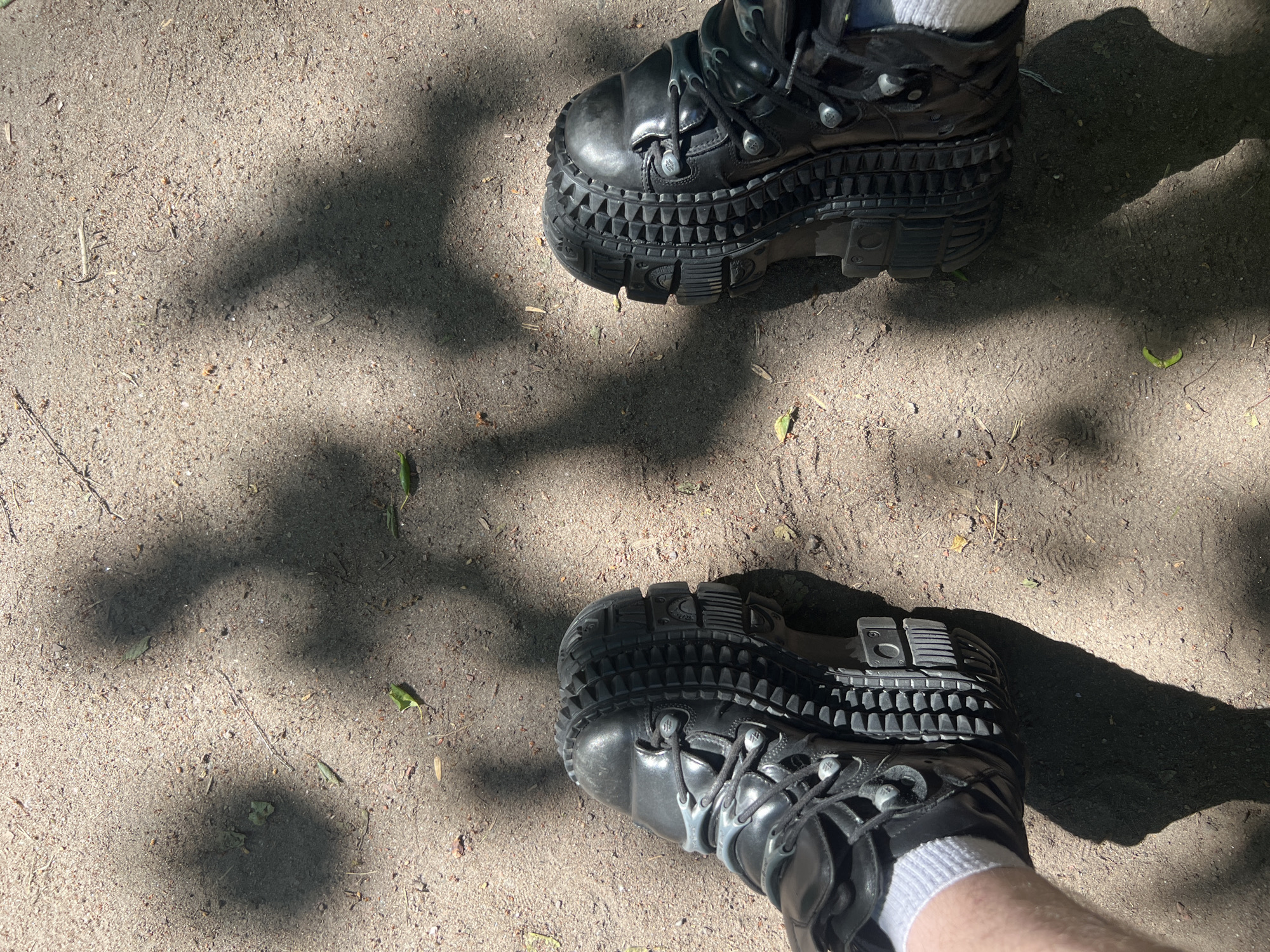 The image presents a ground-level view of a person's feet wearing black, heavy-duty platform boots, standing on a sandy path under bright daylight. Only the lower legs and feet of the person are visible, with white socks peeking out above the boots.
The boots are a prominent feature, characterized by their exceptionally thick, multi-layered platform soles. These soles display an intricate texture, including rows of triangular studs around the perimeter and robust, block-like treads on the bottom. The boots have laces and metallic eyelets or fasteners.
The setting is an outdoor, unpaved area, likely a path or trail, with the ground composed of light brown sand or fine dirt. Scattered on the ground are small bits of natural debris such as dried leaves and tiny twigs. Prominent, dark shadows, likely cast by overhead trees or foliage, create a dappled light effect across the scene, indicating a sunny day. The boots themselves also cast clear shadows.
No specific activity is taking place beyond the person standing on the path. There are no discernible emotions. A small, indistinct logo or text is visible on the side of the sole of the upper boot. The location context provided is Saint Petersburg, Russia.
