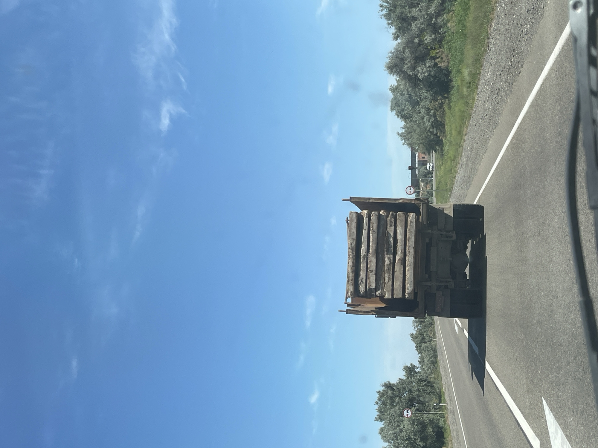 This media file captures a daytime scene in Bakhtemir, Russia, likely from the perspective of a vehicle in motion. The foreground is dominated by the rear of a flatbed truck, heavily laden with what appears to be rectangular, concrete or stone slabs. These slabs are stacked lengthwise, forming a substantial load. The truck itself is dark-colored and partially visible, showing its tires and undercarriage.
The truck is traveling on a paved road with a clear white lane marking. To the right of the road, there is a gravel shoulder and then a dense line of green trees and foliage, suggesting a rural or semi-rural setting. In the distance, along the side of the road, a circular traffic sign with a white background and a red border is visible, featuring two vertical red bars. Further down the road, other structures or vehicles are faintly discernible.
To the left of the truck and road, the scene opens up to a vast, clear blue sky with scattered, wispy white clouds. The bright sky and the quality of light indicate a sunny day. The overall impression is one of a journey along a highway through a natural landscape under favorable weather conditions. There is no visible text other than on the traffic sign, which is not legible enough to decipher any specific meaning. No people are visible in the frame.