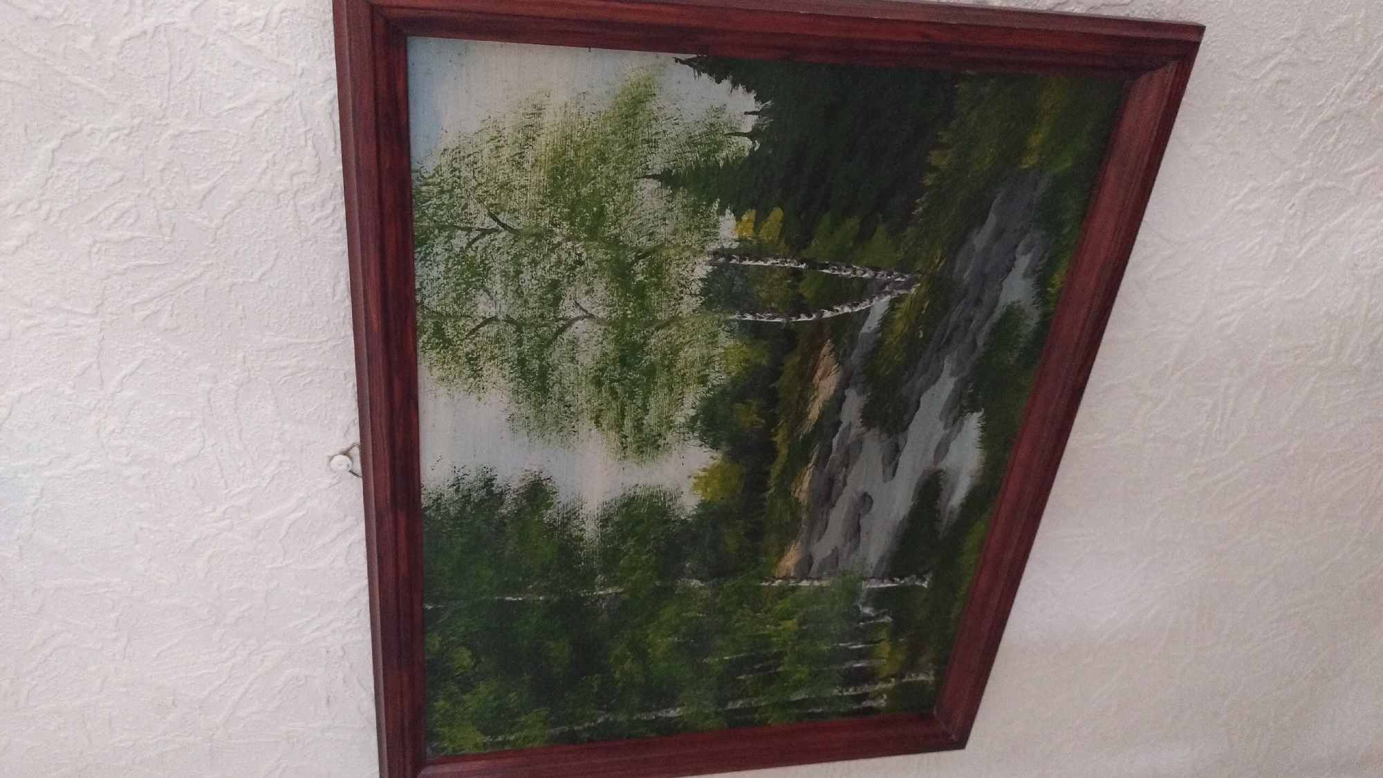 The image shows a framed painting of a forest landscape, possibly a birch grove with a path running through it, hanging on a textured white wall. The painting is in a dark brown wooden frame. The scene depicts several birch trees with white bark and green foliage, along with other trees and shrubs in shades of green and dark green. A path, which appears to be made of stones or a rough surface, winds through the trees. The sky in the background is a light, overcast gray. There are no people visible in the painting or the immediate surroundings. The painting is hanging on a wall in what appears to be an indoor setting. The location in the city of Tukums, Latvia, is not identifiable from the visual information provided in the image.