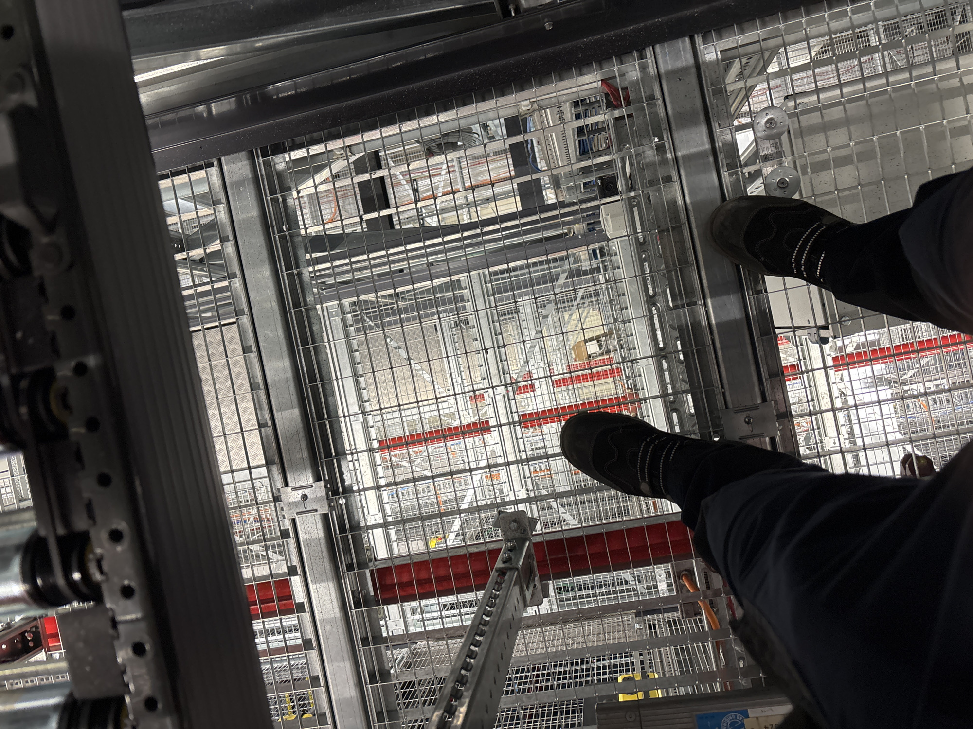 The image is a first-person perspective of someone standing on a metal grate platform, looking down into a complex industrial or logistical facility. The individual is wearing dark work pants and black shoes with white stitching. The platform is elevated, with multiple levels of similar grated flooring and structural metalwork visible below. Some of the lower levels feature red vertical elements and various racks or shelving units. The overall environment appears to be a warehouse or distribution center. The angle of the shot suggests the person is standing near an edge, with a significant drop below. The location is identified as Mannheim, Germany, implying this facility is within that city.