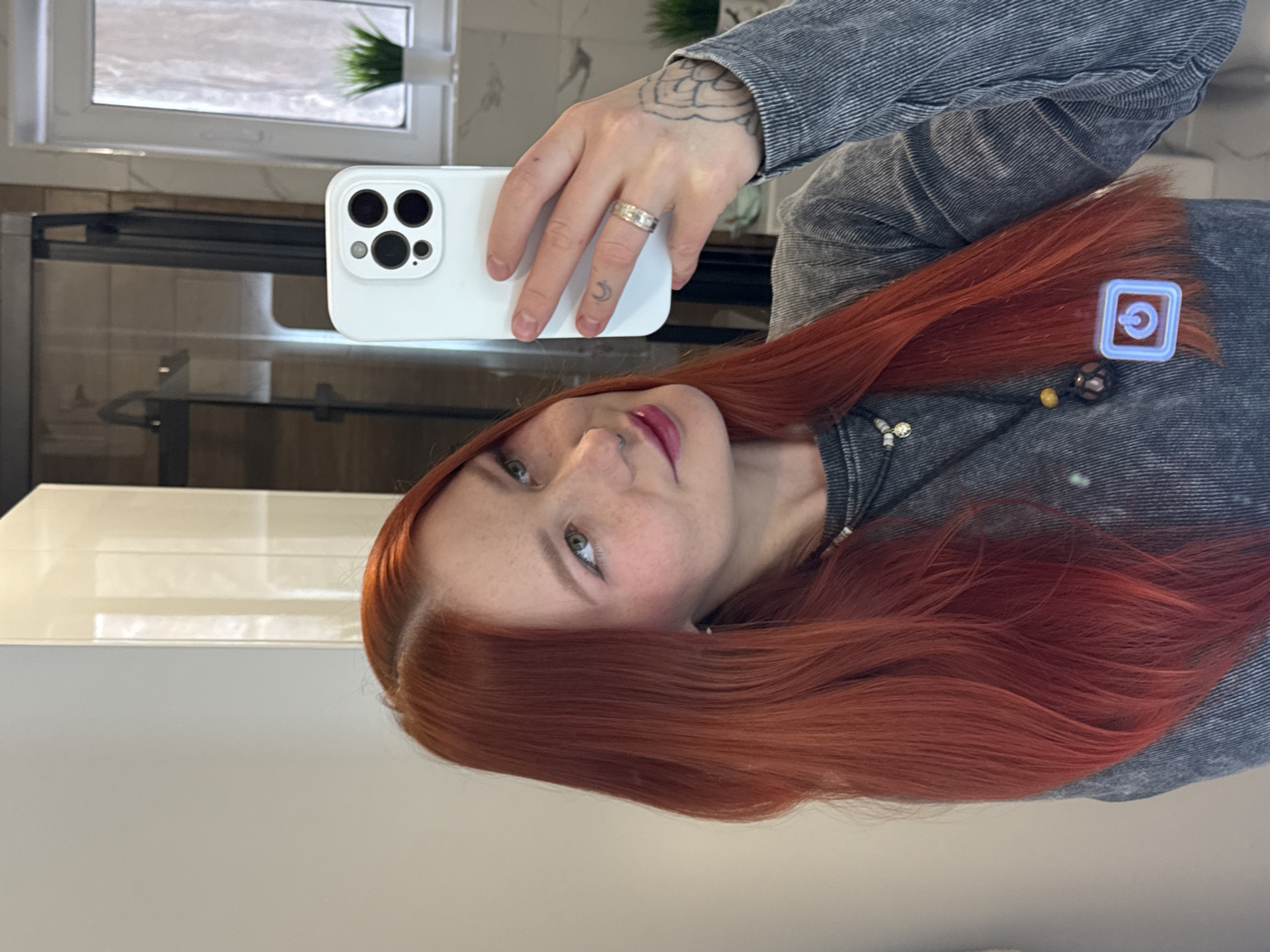 An adult female with long, straight, reddish-orange hair, light skin, and freckles is captured in a mirror selfie within what appears to be a bathroom in Rostov-on-Don, Russia. She has light-colored eyes and wears a silver nose ring in her right nostril. Her lips are adorned with a reddish-pink cosmetic. She is dressed in a grey, textured long-sleeved top, accessorized with two necklaces: a shorter one with a white shell bead and silver charm, and a longer one featuring dark beads and a larger, dark pendant. A neon blue-outlined power button icon is digitally overlaid on her chest.
Her right hand, displaying a silver-colored ring on the ring finger and a crescent moon tattoo on the middle finger, holds a white smartphone (identified as an iPhone 14 Pro/Pro Max due to its camera array) towards the mirror to take the photograph. A floral/leafy tattoo is visible on the back of her right hand/wrist.
The reflected background reveals a modern bathroom setting. To the left, a plain light-colored wall or cabinet is visible. Behind the phone, a dark-framed shower enclosure with a dark shower fixture is reflected. To the right, a white-framed window with an obscured or frosted view, along with a small potted artificial green plant on a ledge below it, is present. The wall surrounding the window appears to be tiled with light-colored, possibly marble-patterned, rectangular tiles.