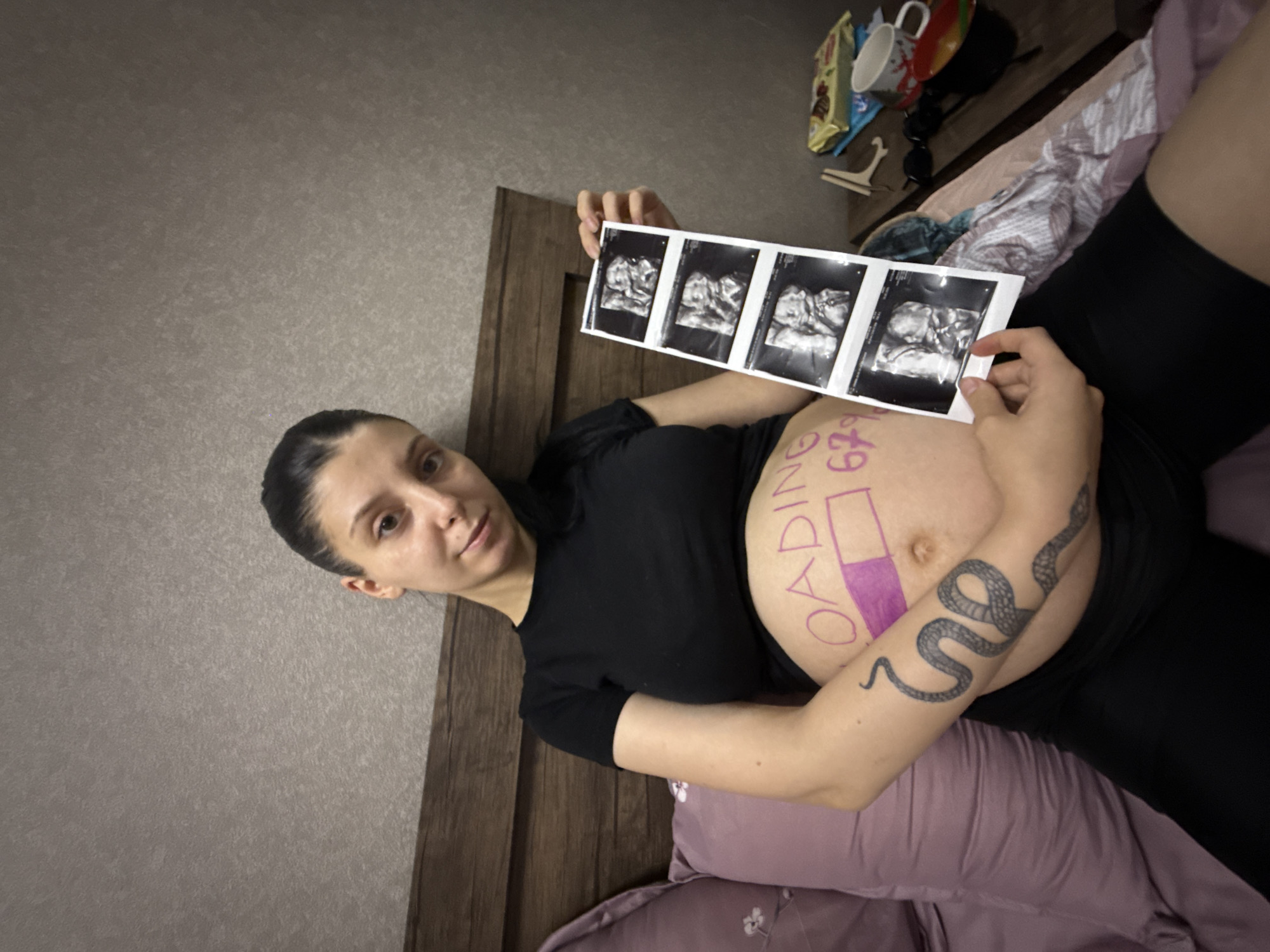 A woman is lying on her side in bed, holding up a strip of ultrasound images. She is wearing a black t-shirt and black leggings. Her pregnant belly is visible, with the word "LOADING" written on it in purple marker, along with a progress bar. She has a snake tattoo on her arm. The background appears to be a bedroom with a textured grey wall and a wooden headboard. There are some items on the bedside table, including a mug, a packet of snacks, and sunglasses. The lighting suggests it is daytime. The overall mood is one of anticipation and joy associated with pregnancy. The location context is Tbilisi, Georgia.