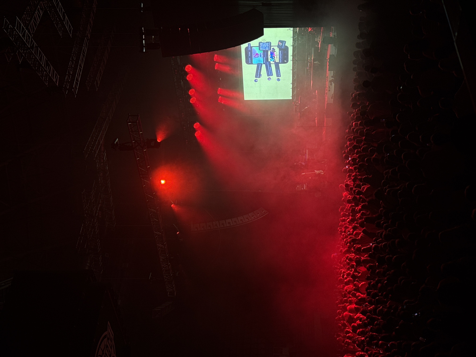The image depicts a large, dark concert hall filled with a dense crowd of people. Red stage lighting illuminates the scene, creating a smoky atmosphere. A large screen is visible on the stage, displaying an image of a smartphone with various notifications and a QR code. Several speakers are mounted on the stage and suspended from the ceiling. The overall impression is that of a live music performance taking place in a venue. There is no visible indication of the city of Krasnoyarsk, Russia.