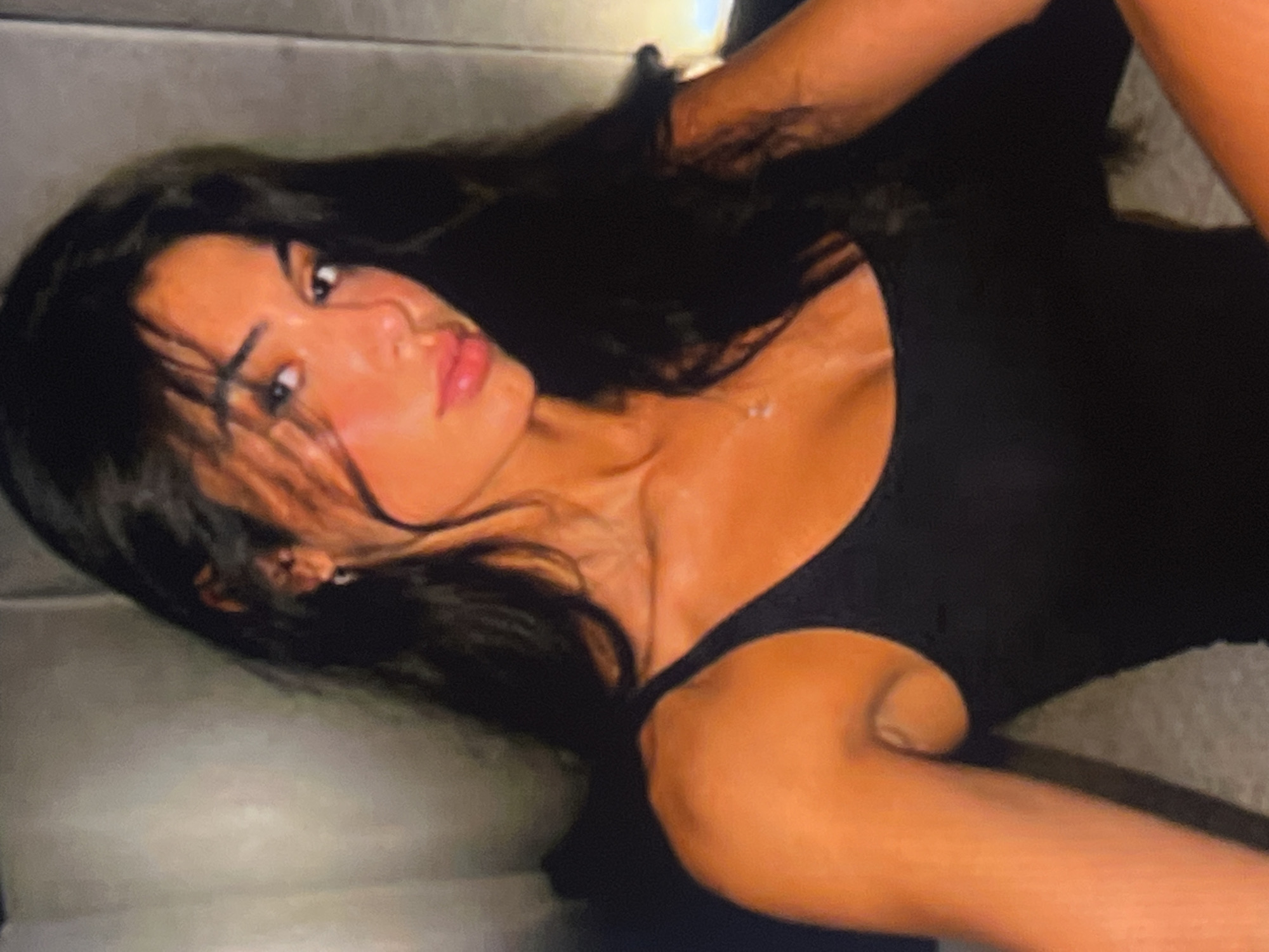 A medium shot shows a young woman with long, dark, wavy hair and dark eyes, wearing a black tank top. She has tanned skin and is looking at the camera. Her left arm is visible, extended towards the viewer. The background appears to be an indoor setting with textured walls. The lighting is warm and casts a glow on her skin.