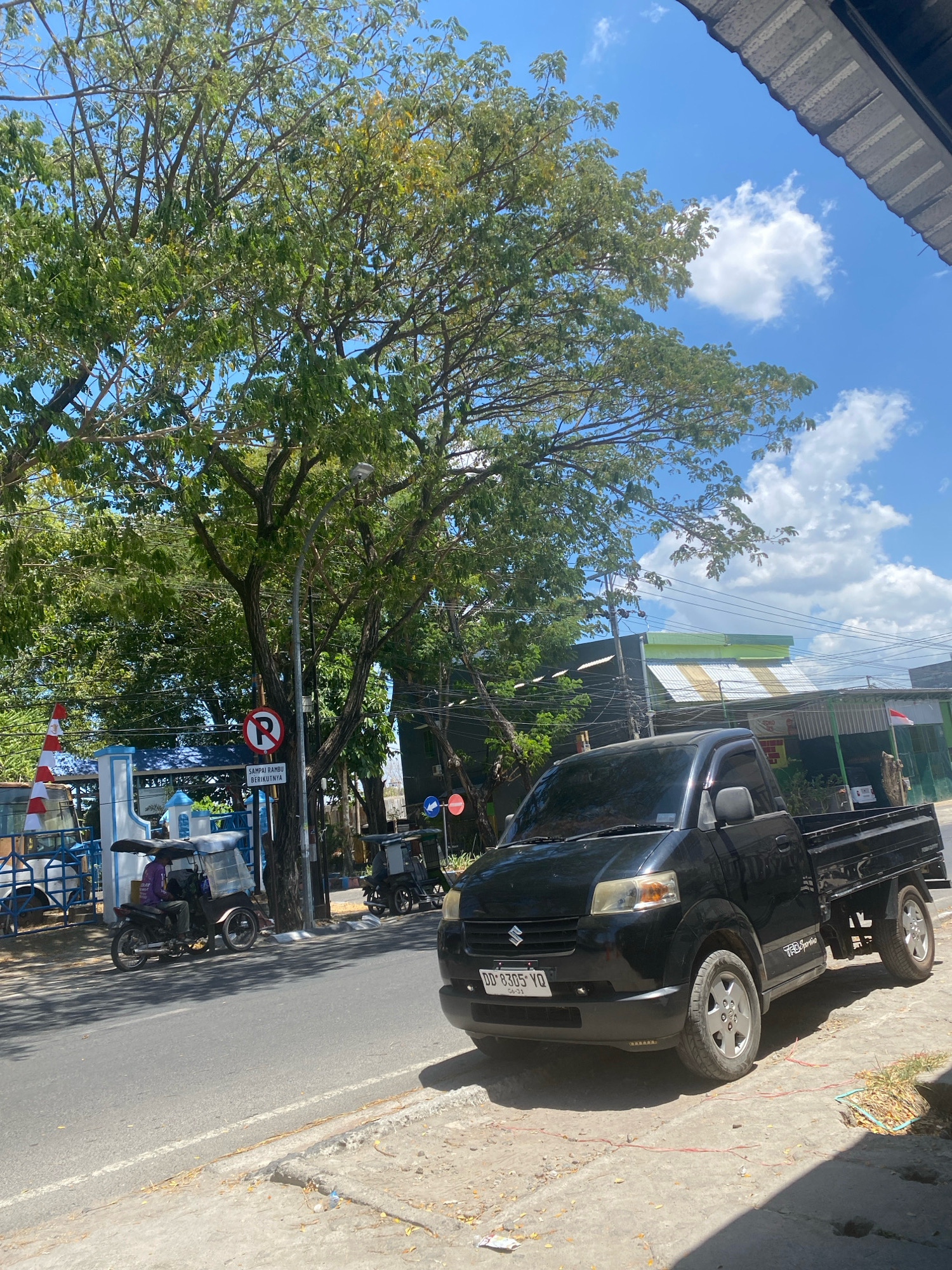 This is a full-frame, eye-level, daytime, outdoor shot of a street scene in Kalumeme, Indonesia. The focal point is a black Suzuki Carry pickup truck parked on the right side of the road. The truck has a license plate that reads "DD 8305 VO" and has "TRD Sportivo" branding on its side. To the left of the truck, a man wearing a purple shirt is riding a three-wheeled motorcycle. Further left, there is a white and red traffic cone and a blue and white structure with railings. A "no parking" sign is visible on a pole near a large tree. Behind the truck and other structures, there are buildings with corrugated metal roofs. The sky is blue with scattered white clouds. The road is asphalt, and the sidewalk is made of concrete and dirt.