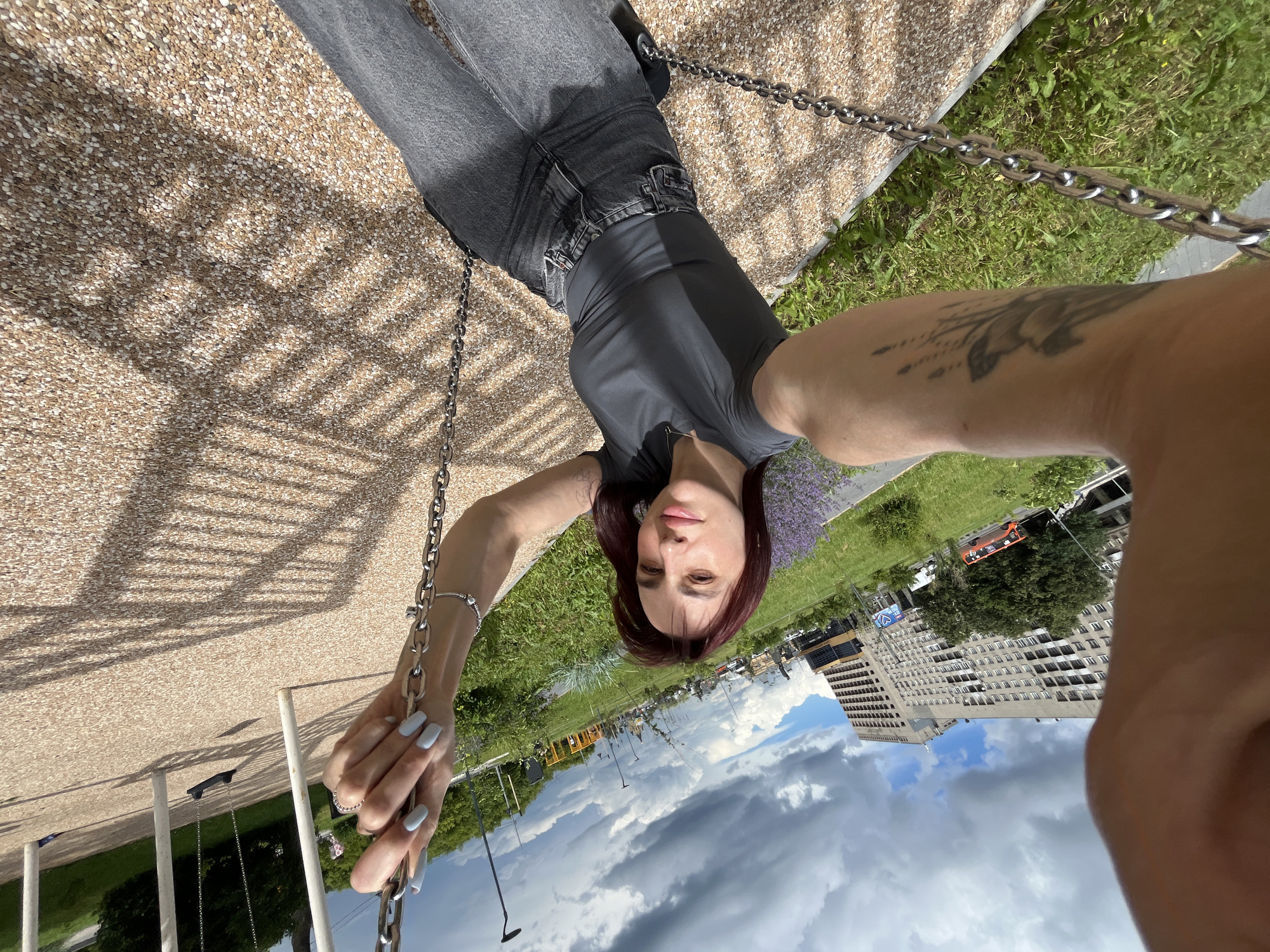 A young woman with dark red hair is taking a selfie while sitting on a swing in an outdoor park. She is wearing a dark grey t-shirt and dark grey jeans. Her left arm, which is holding the swing chain, is visible and has a tattoo of a floral design. The swing has a metal chain and appears to be made of black rubber. The ground of the park is covered in small, light-colored pebbles. In the background, there is a grassy area with some purple flowers, trees, a tall building, and other playground equipment. The sky is partly cloudy. The woman's fingernails are painted a light blue color, and she is wearing a silver ring and bracelet. The image appears to be taken on a sunny day. The presence of the tall building and the overall urban park setting suggest this could be a location in the city of Nizhny Novgorod, Russia, possibly a newly developed recreational area or a public space with modern amenities.
