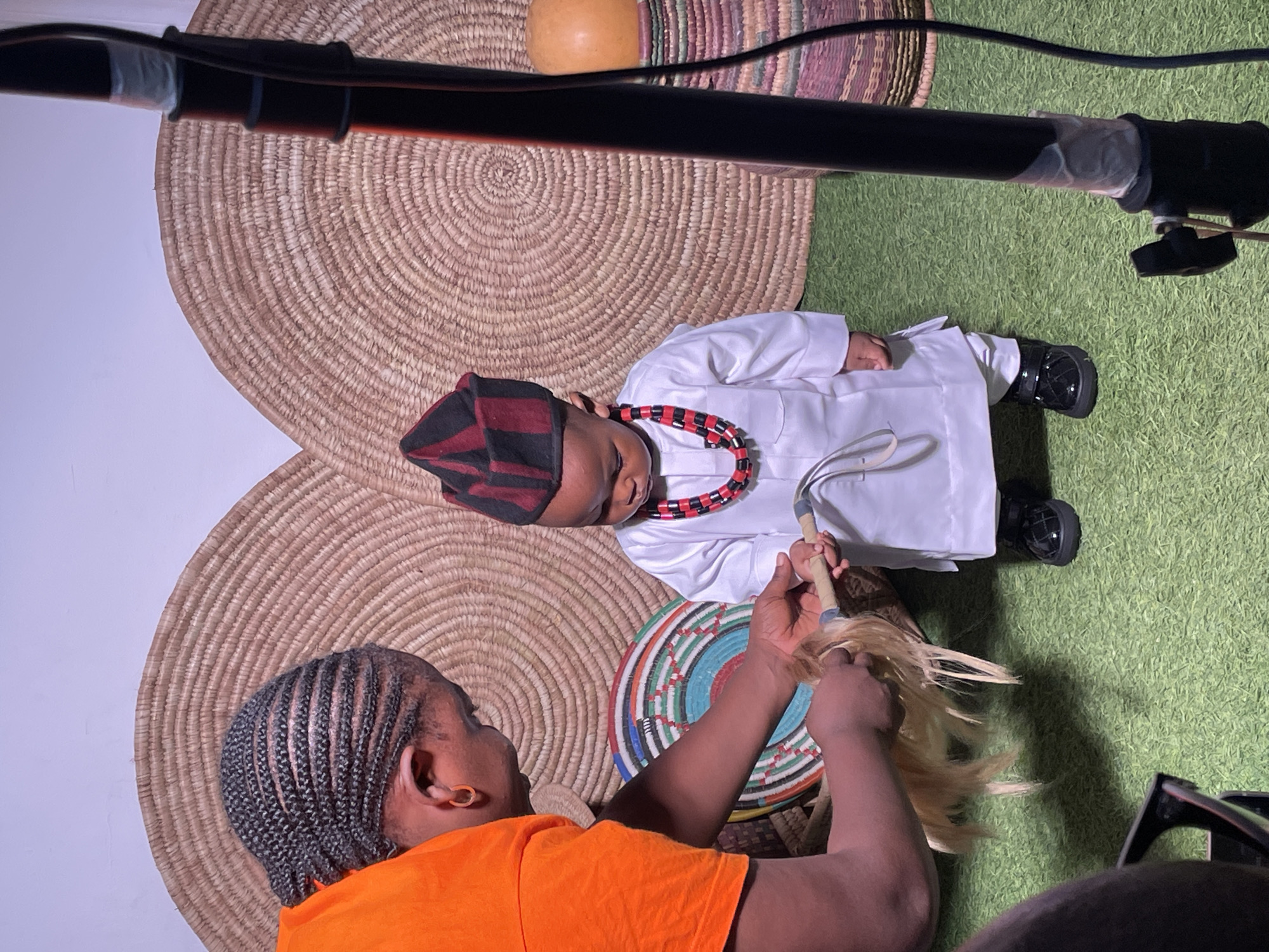 In Jos, Nigeria, a young child is being dressed for a photoshoot by an adult. The child, wearing a white traditional outfit and a striped cap, stands on a green, artificial turf floor. A beaded necklace of red and black is around the child's neck. The adult, dressed in an orange t-shirt, is adjusting a piece of blond hair-like material attached to a wooden stick held by the child. The background features two large, woven circular mats, and to the right, a woven cylindrical object with a rounded top is visible. A black lighting stand is also present on the right side of the frame. The scene appears to be an indoor studio setup designed to evoke a cultural aesthetic.
