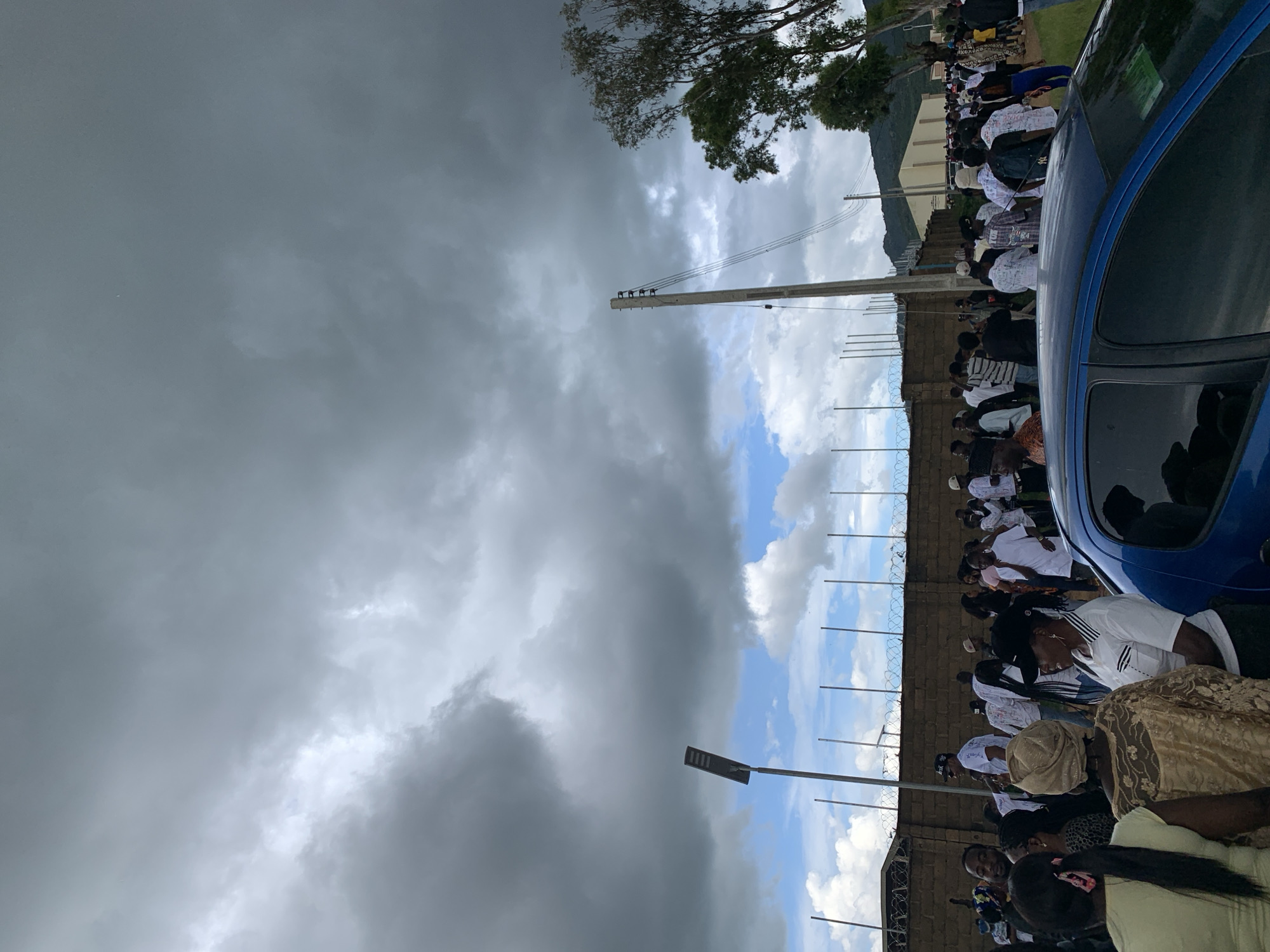 The image depicts a large gathering of people outdoors, with a dark, cloudy sky overhead, suggesting an impending storm. The crowd is composed of individuals of various ages and genders, dressed in casual and traditional attire. Many are wearing white clothing. In the foreground, the rear of a blue car is visible. A tall street light with a rectangular panel stands to the left of center, and a utility pole with wires stretches across the frame to the right. A brick wall topped with barbed wire runs horizontally across the middle ground, separating the crowd from what appears to be an open area. In the distance, trees and buildings are visible, with a mountain range forming the backdrop. The overall impression is a scene of social gathering or an event taking place in an urban or semi-urban environment, possibly in Yelwa, Nigeria.