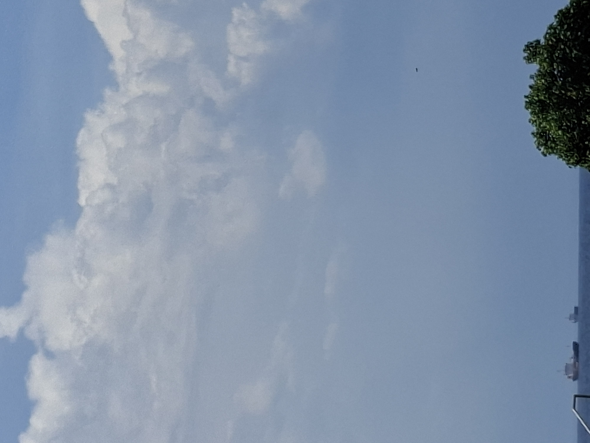 The image shows a daytime view of the sky, partially obscured by a large, white cloud formation on the left side. A tree with dense green foliage is visible on the right edge of the image. In the lower right corner, a building or structure, perhaps a multi-story hotel, is faintly discernible. The sky is a pale blue. Given the location of Kuta Selatan in Indonesia, which is on a tropical island, the scene could be a coastal area near the city, although nothing in this image verifies the exact location.