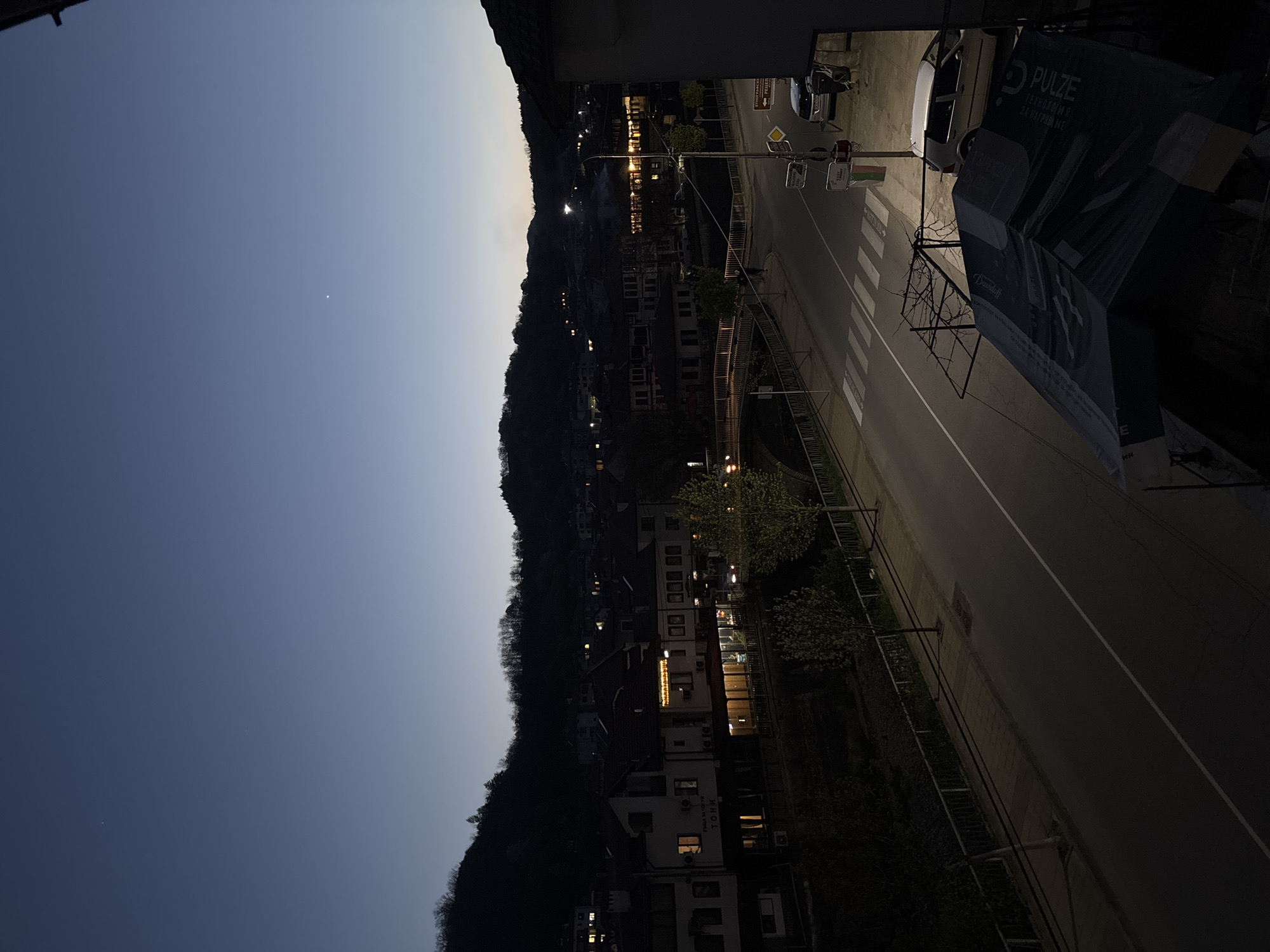 The image captures a panoramic view of a town nestled in a valley, likely Zlatograd, Bulgaria, during dusk or early night, taken from an elevated perspective. The sky transitions from a deep indigo at the upper left to a lighter, paler blue-grey near the horizon line on the right, where a solitary bright star or planet is visible. Dark, rolling hills or mountains silhouette against the fading light, framing the town below.
A paved road with white lane markings, including a solid line, dashed center lines, and a pedestrian crosswalk, runs through the scene. Streetlights illuminate sections of the road, casting long glows. Several buildings line both sides of the road, many with warm, illuminated windows suggesting activity within. On the left side, closer to the foreground, a multi-story building with "КУКЛА ЗА ГОСТИ ТОРИ" (Guest House TORI) visible on its facade stands.
A white car is parked on the right side of the road, near a large dark banner hanging from an overhead structure. This banner advertises "PULZE / ТЕХНОЛОГИЯ ЗА ГРАДА НИ" (PULZE / TECHNOLOGY FOR OUR CITY) and also features the brand "Davidoff." Traffic lights and a streetlamp are visible along the road, accompanied by railings or fences. The overall scene is quiet, with no visible human activity, conveying a peaceful evening atmosphere in the mountainous town.
